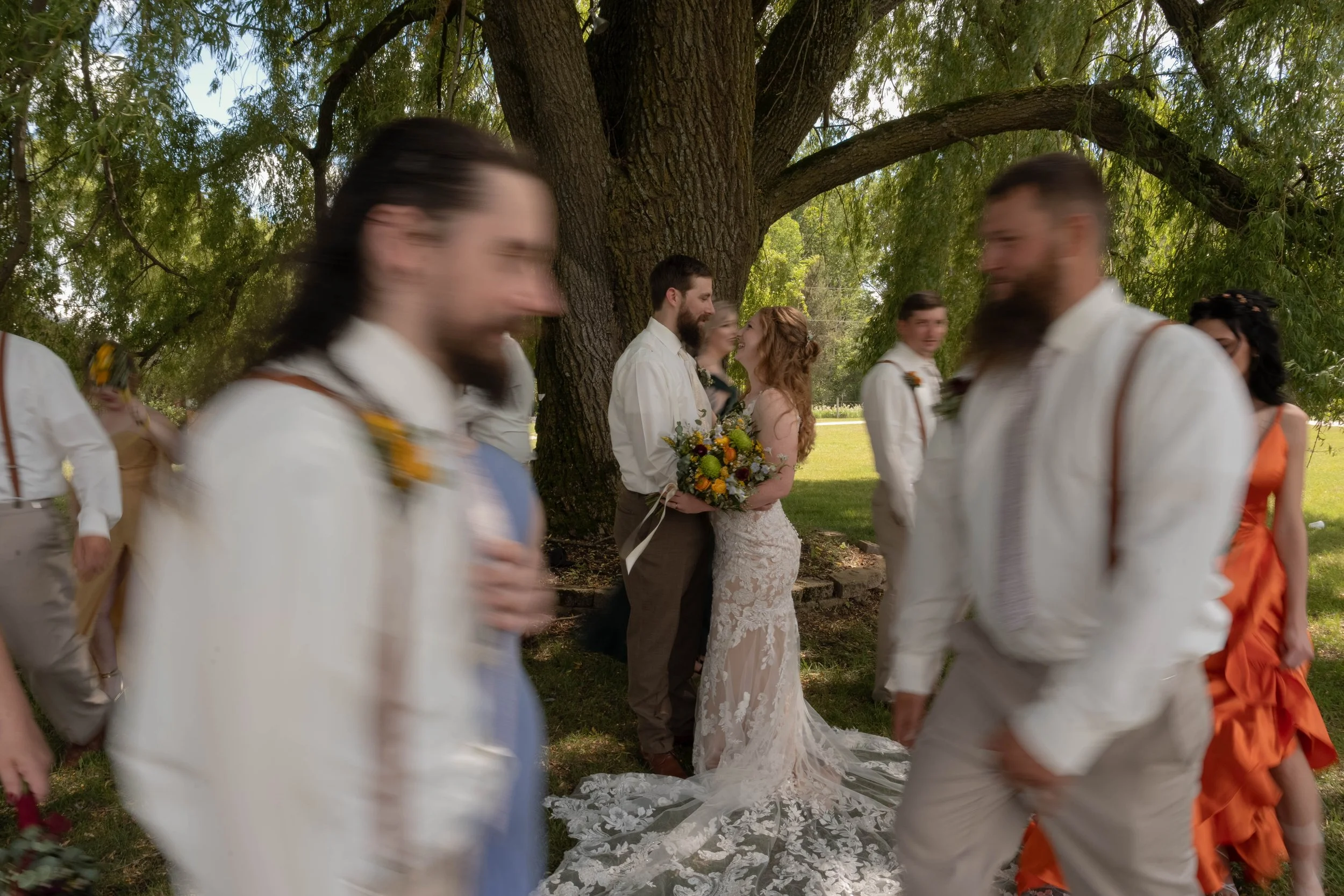 A bride and groom standing closely and smiling under a large tree, surrounded by guests dressed in white and colorful outfits, outdoors during daytime.