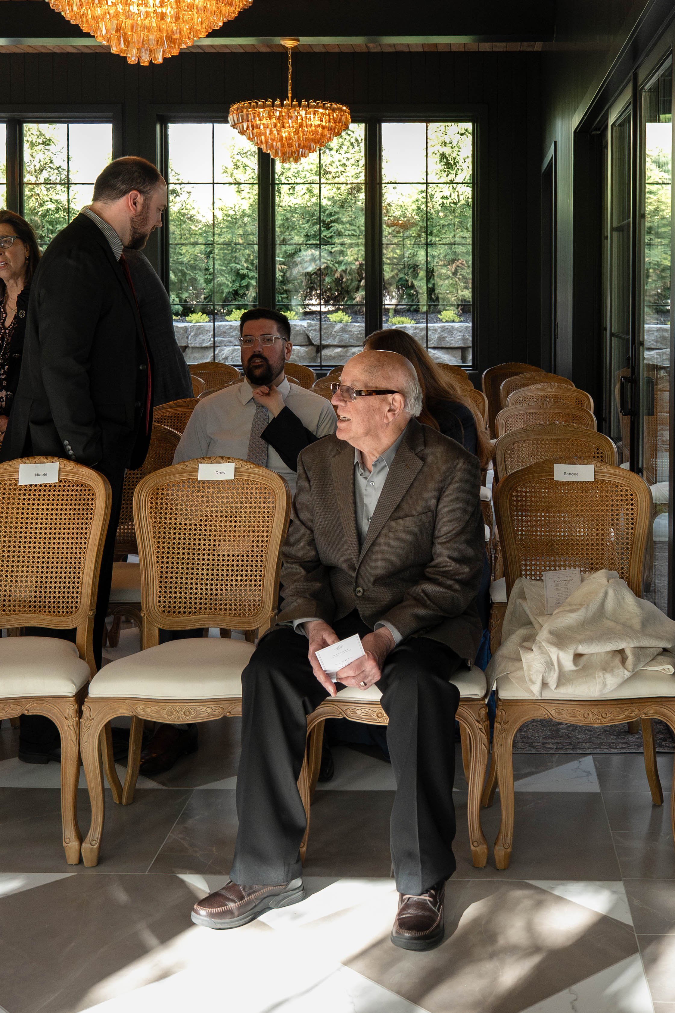 A group of people at a formal event, sitting and standing in a room with large windows, greenery outside, a chandelier, and chairs with name tags.