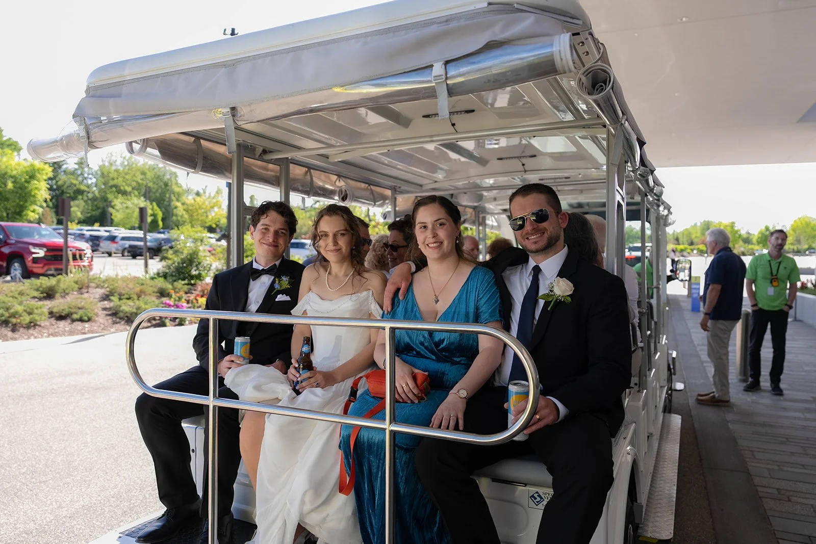 Group of four people sitting in a golf cart during a celebration or wedding, smiling at the camera. The group includes a groom in a tuxedo, a bride in a wedding dress, and two women dressed nicely, one in a blue dress and one in black suit with sungl