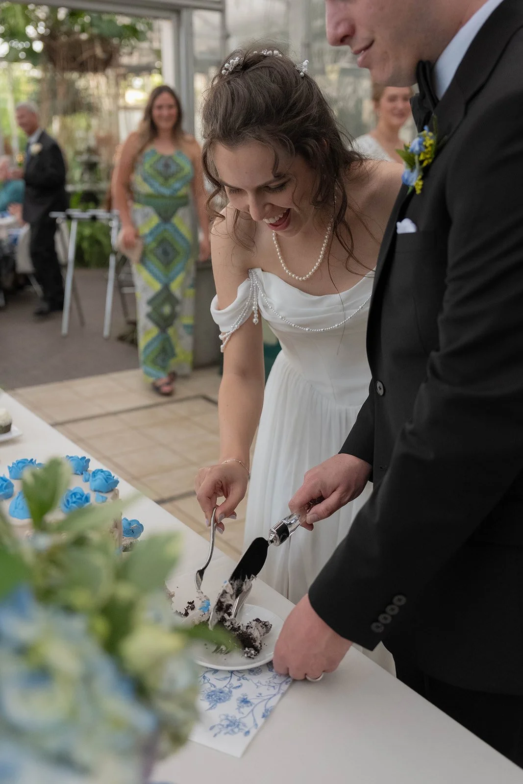 A bride and groom cutting a wedding cake together during their reception, with guests in the background.