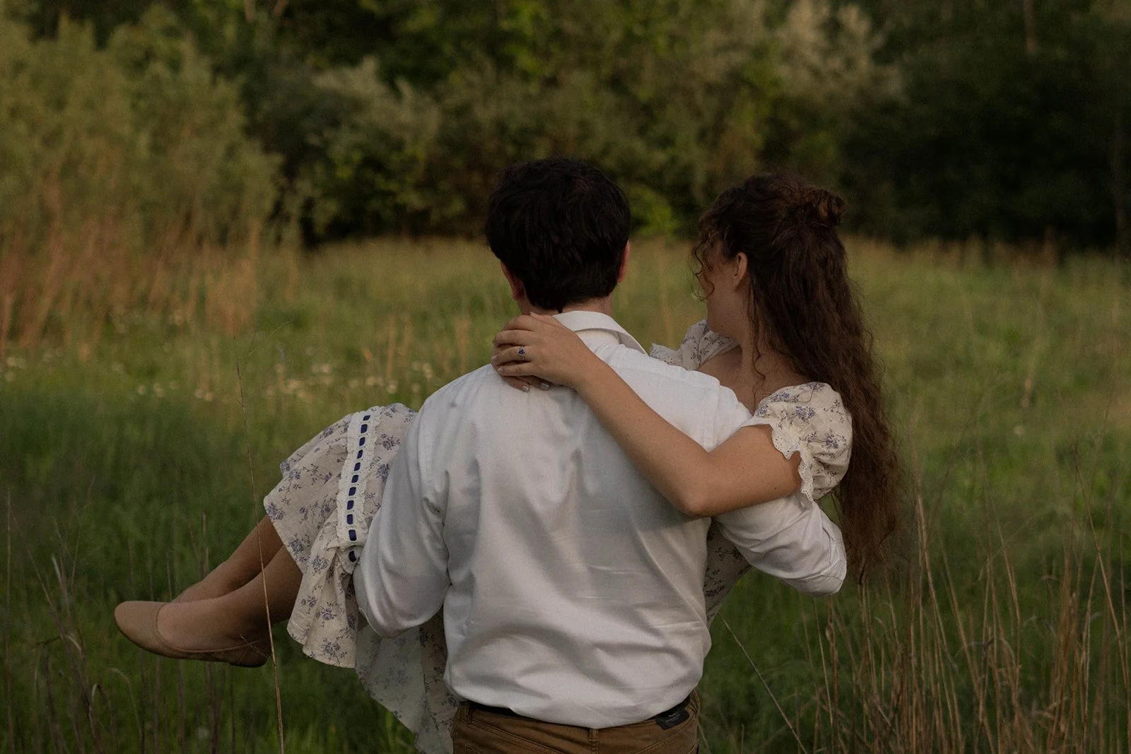 A couple embracing outdoors in a grassy field with trees in the background.