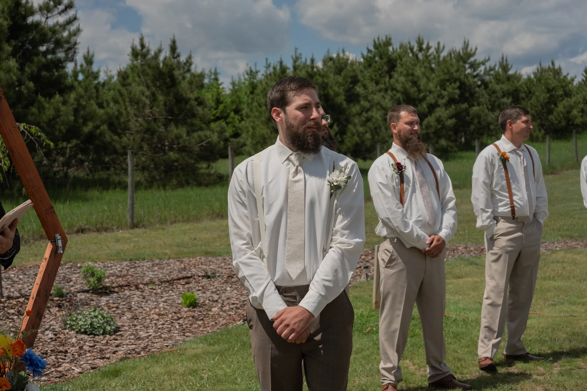 Three men standing outdoors during a wedding ceremony, dressed in formal attire with flower boutonnieres, in front of a backdrop of green trees and a partly cloudy sky.