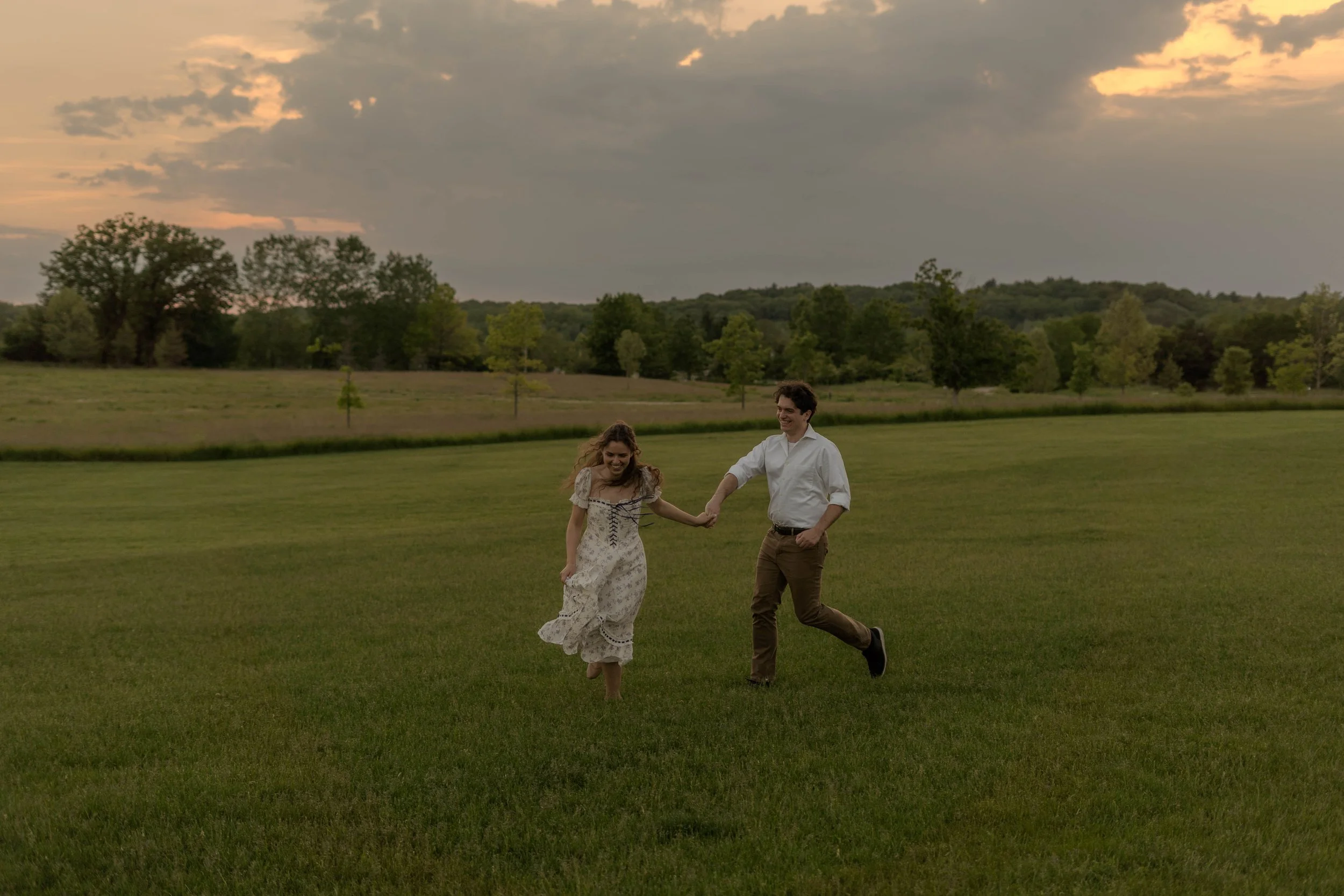A smiling young man and woman holding hands and running on a grassy field during sunset, with trees and a cloudy sky in the background.