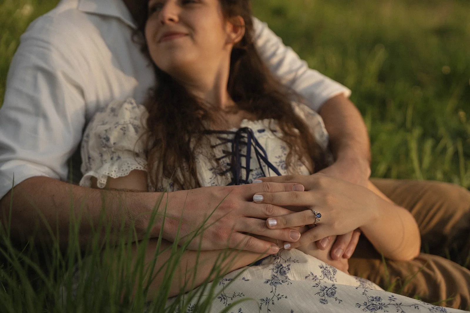 A woman with long, wavy hair lying in the grass, smiling, with a man embracing her from behind. She is wearing a white dress with lace and dark string details, and she has a ring on her left hand.