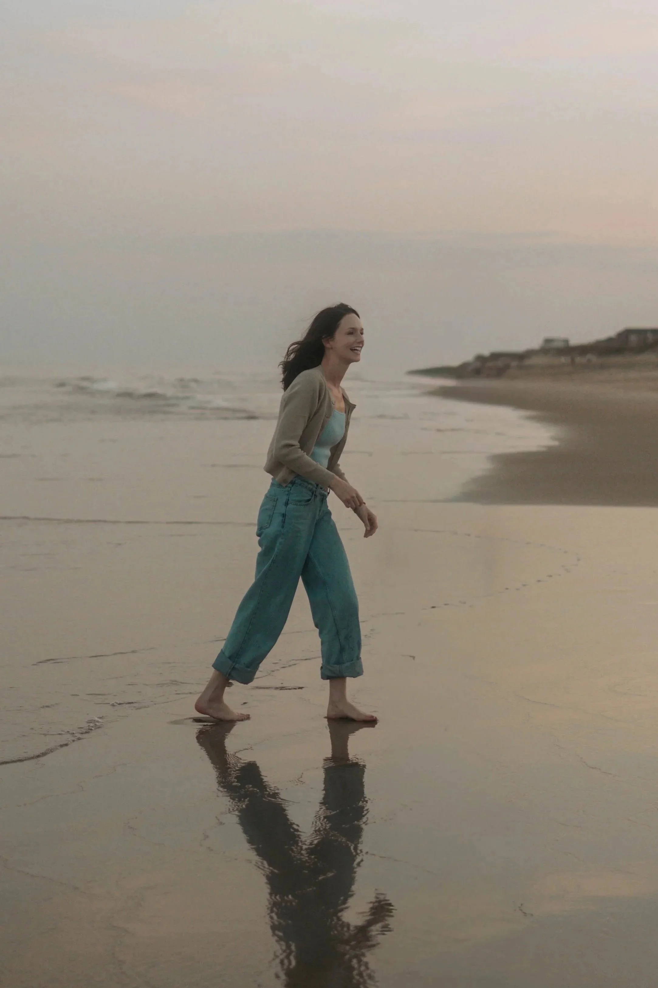 A woman walking barefoot on the wet sand at the beach during sunset, smiling and enjoying the moment.