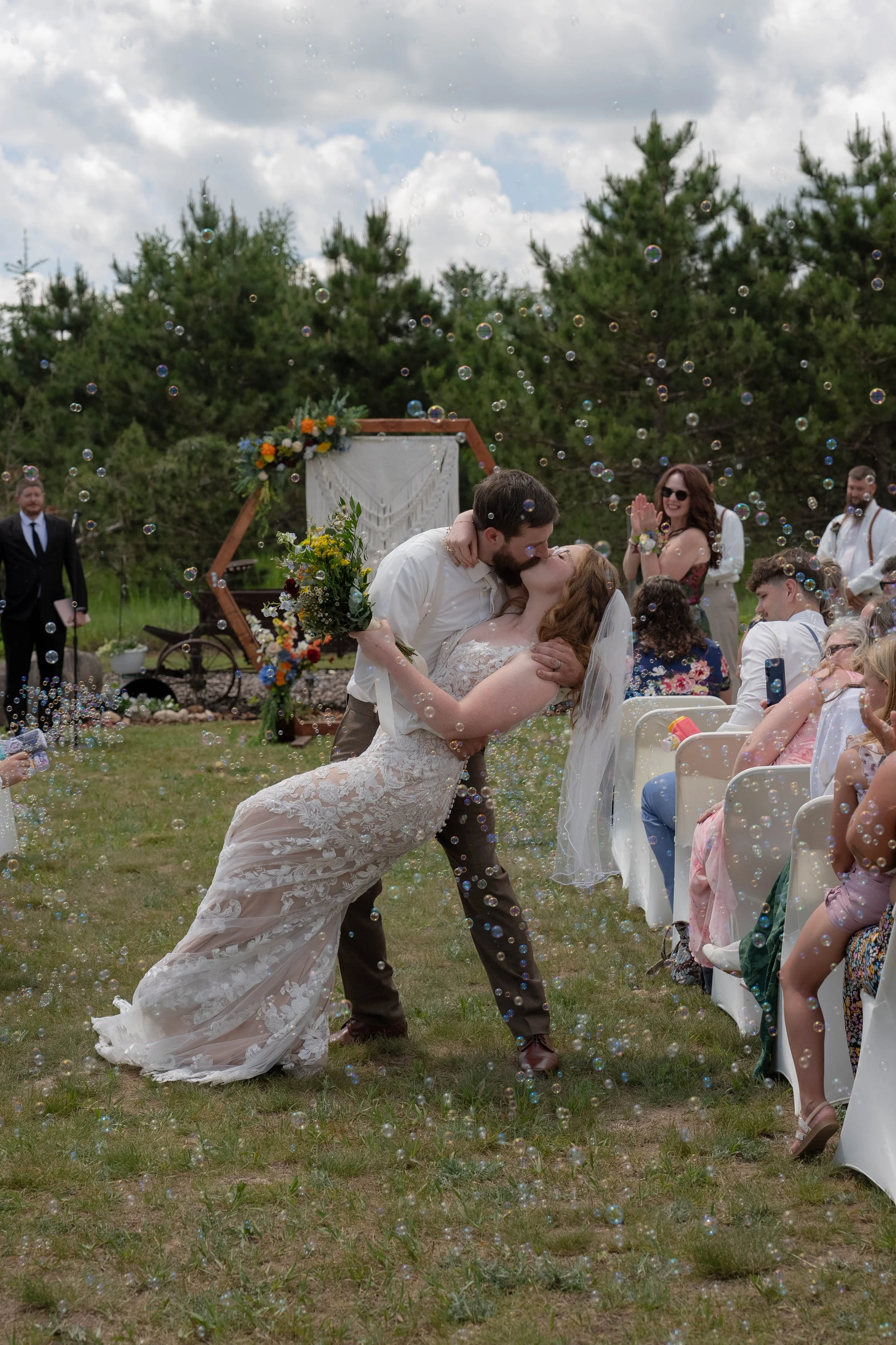 A couple shares a kiss at an outdoor wedding ceremony, with bubbles floating around and guests seated nearby.