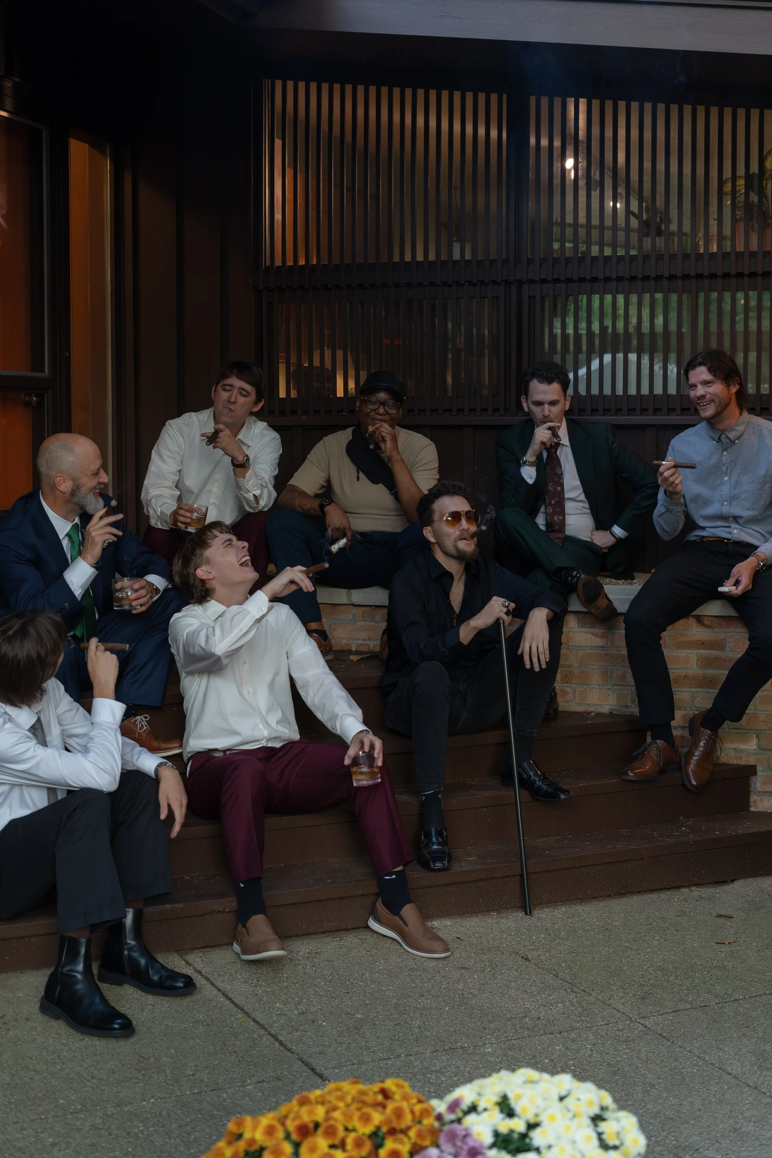 A group of people sitting and standing on a wooden porch, enjoying a social gathering with flowers in the foreground and a dark building wall with windows behind them.