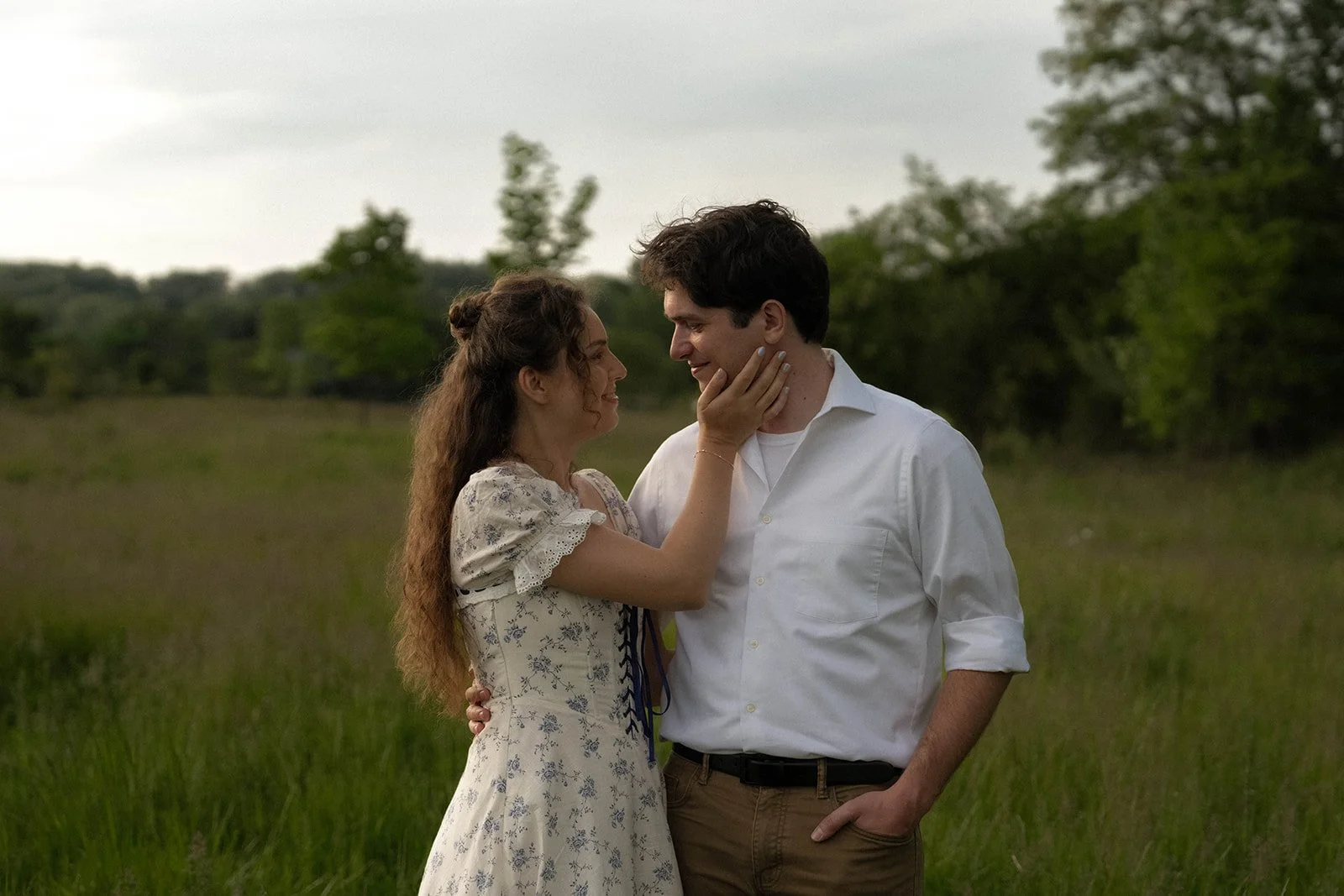 A young woman and man standing close together in a grassy field, smiling and gazing into each other's eyes. The woman gently touches the man's face while holding him close with her other arm, and they appear to be in a romantic moment.