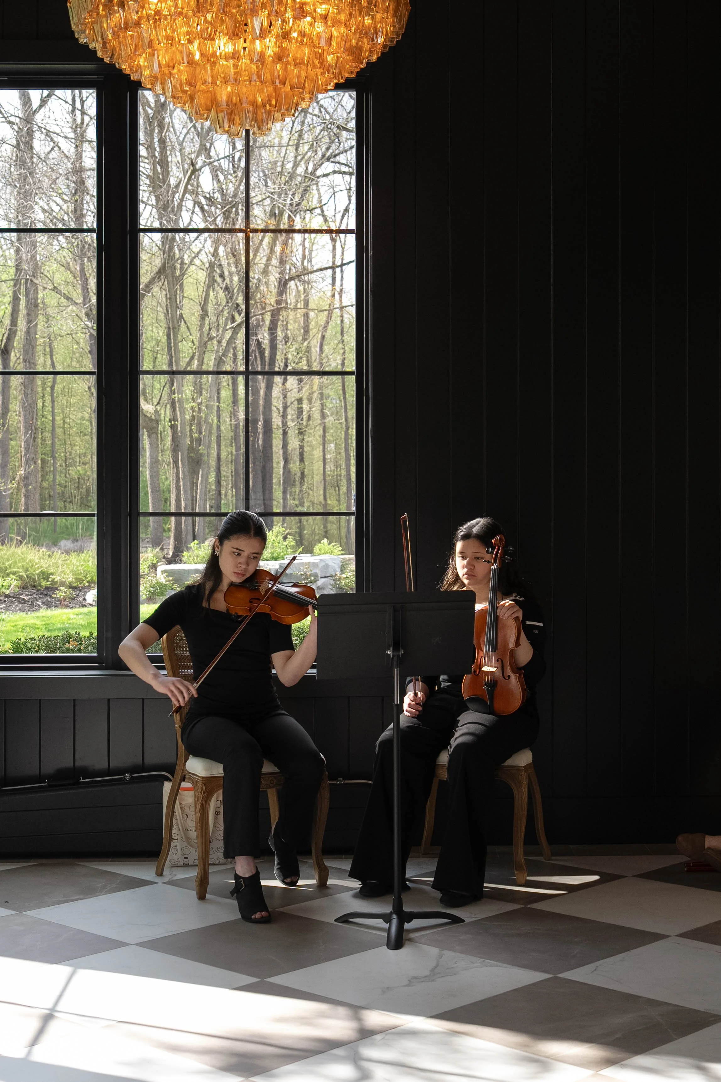Two women playing violins indoors near large windows with a view of trees. One woman is seated on a wooden chair, and the other woman is seated on a cushioned chair. A music stand with sheet music is in front of them. An elegant chandelier hangs from