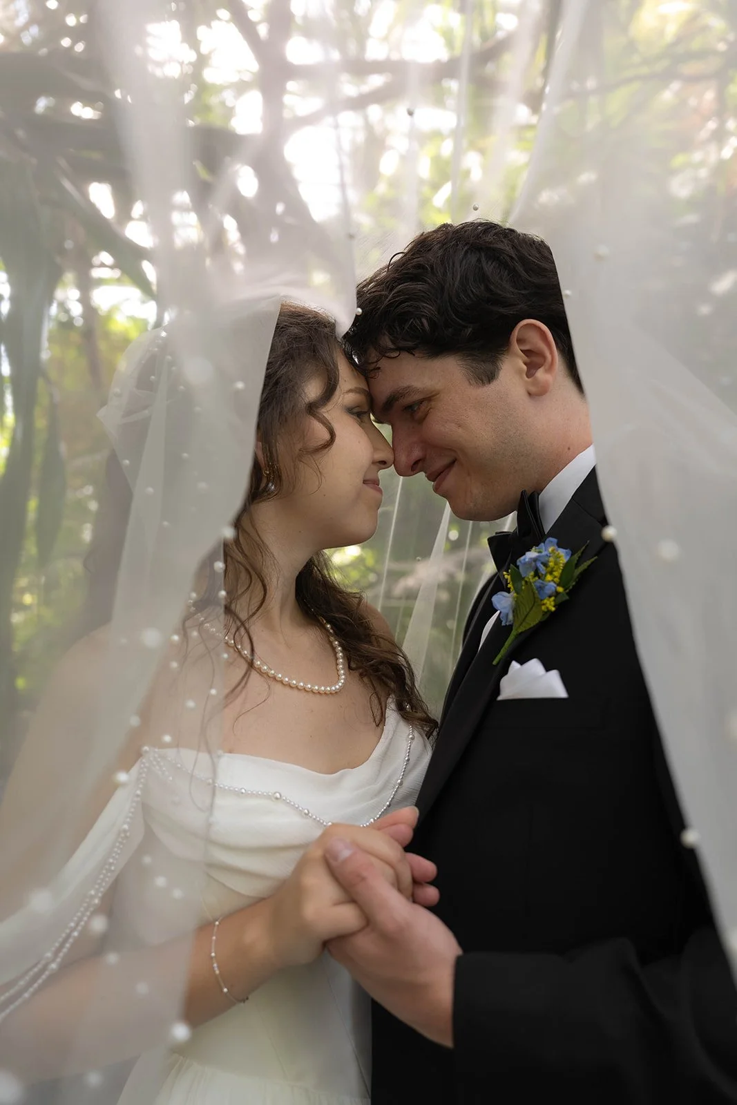 A bride and groom face closely under a sheer veil, holding hands, outdoors with trees in the background.