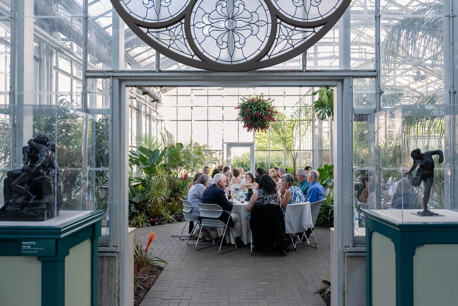 People seated at round tables in a conservatory filled with lush green plants, with glass walls and ceiling, enjoying a gathering.