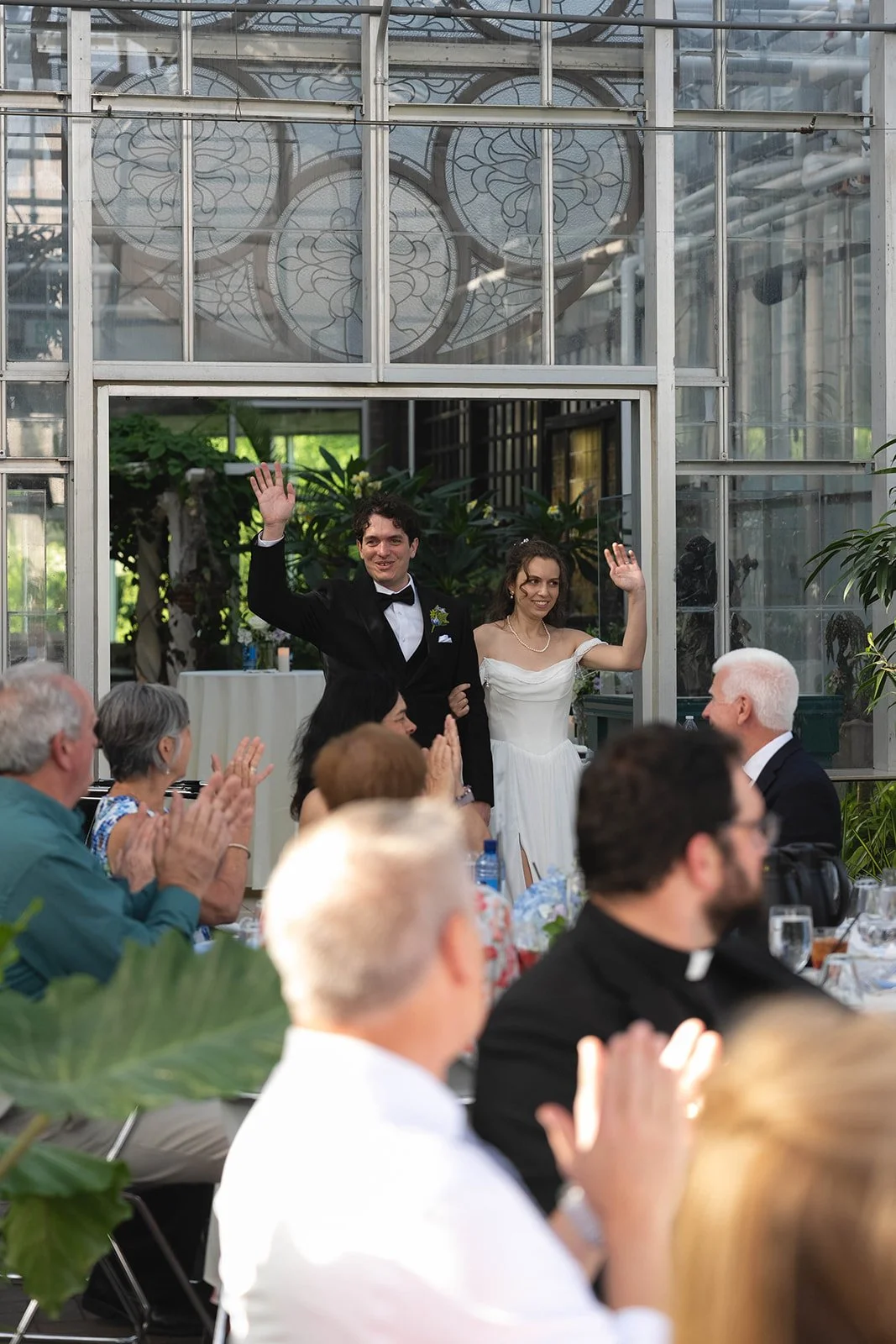 A newly married couple waving at guests during their wedding reception in a greenhouse or conservatory setting, with guests clapping and smiling at the couple.