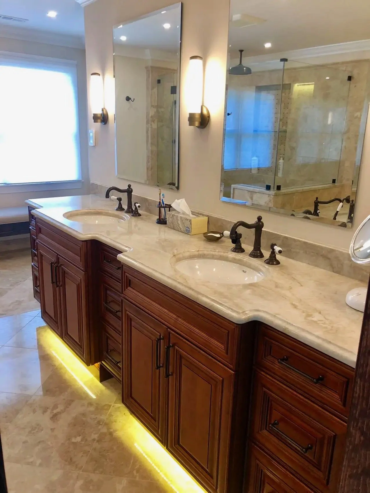 A bathroom vanity with dual sinks, wood cabinets, marble countertop, and large mirror. In the background, a glass-enclosed shower with beige tile walls and ceiling-mounted showerhead. Light fixtures and a window provide illumination.