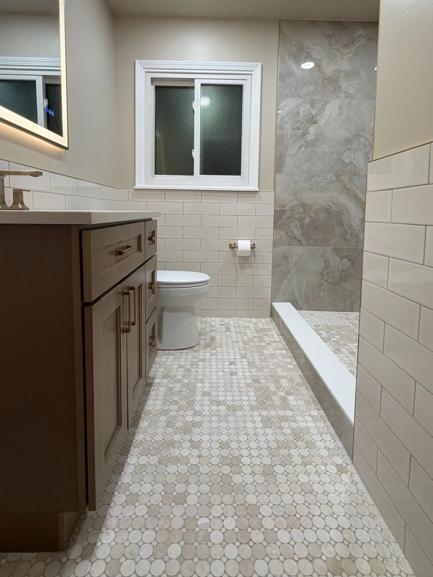 A modern bathroom featuring a small vanity with dark wood cabinets, a white countertop, a toilet, a frosted window, a mosaic tile shower with a wood-look accent tile wall, and a beige hexagonal tile floor.
