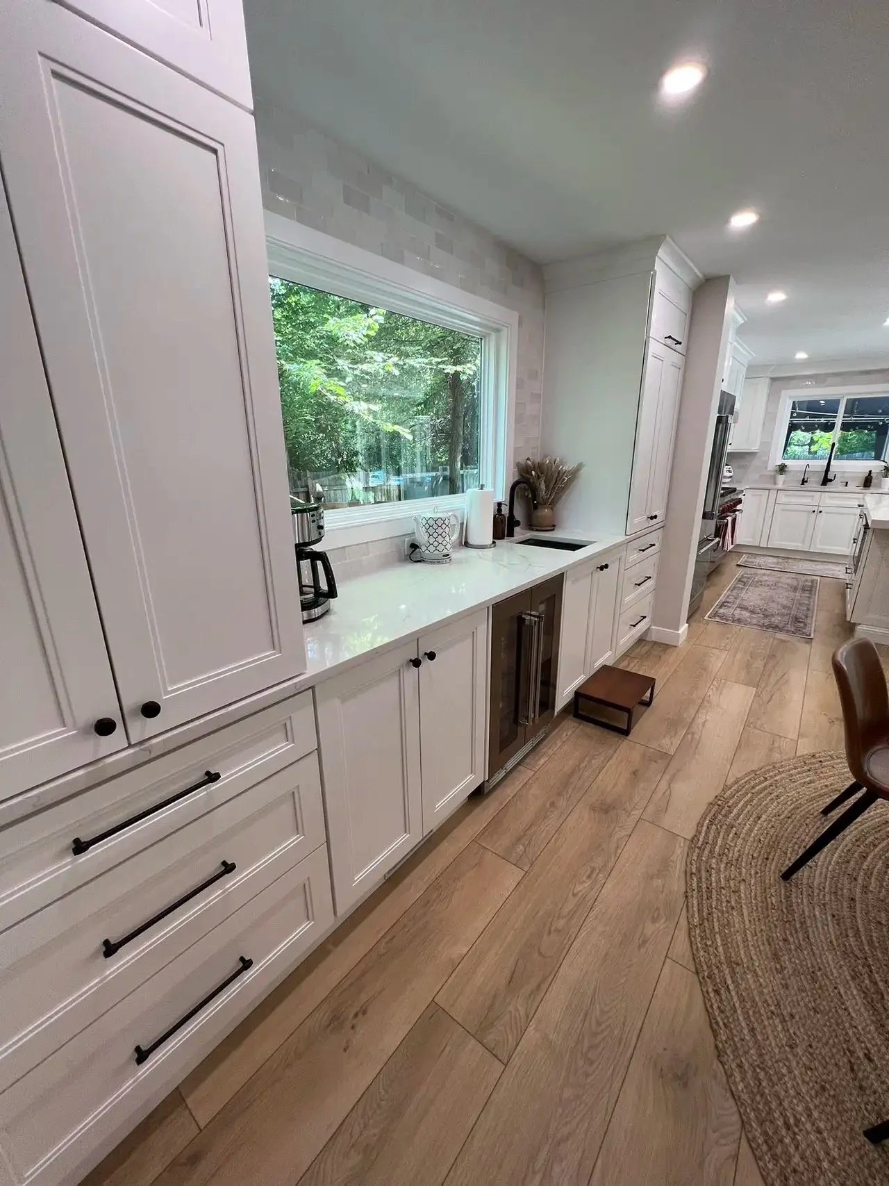 Modern kitchen with white cabinets, a large window revealing green trees outside, and wooden flooring. The countertop has a coffee maker, paper towel holder, and decorative plant. There is a small stool near the cabinets and a rug on the floor.