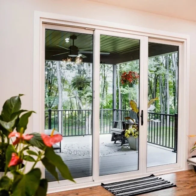View of a screened porch from inside a house, featuring a sliding glass door, a hanging flower basket, potted plants, a rocking chair, and trees in the background.