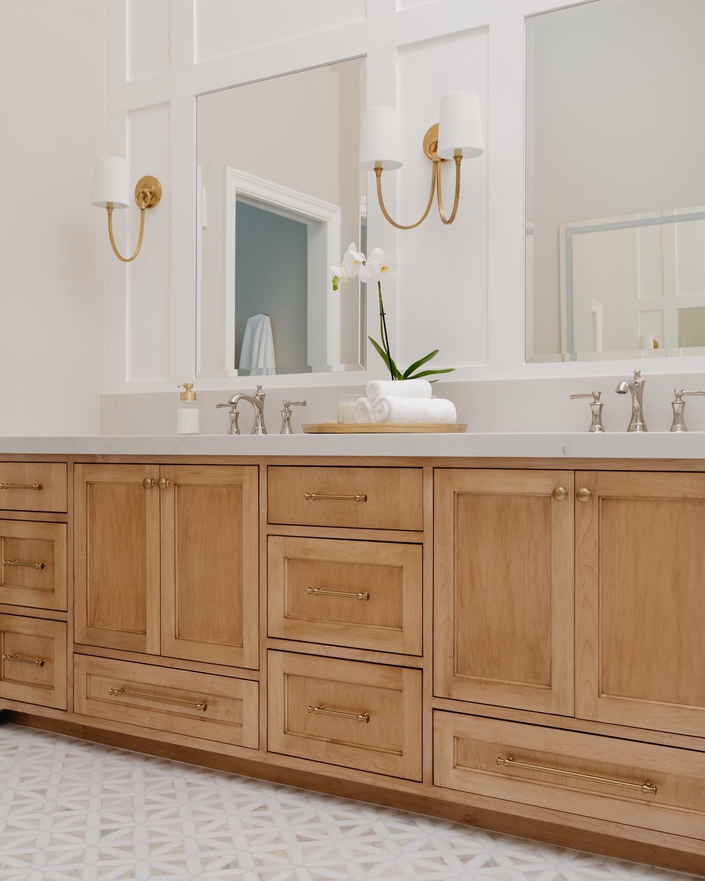 Bathroom vanity with wooden cabinets, white countertop, and silver faucets, decorated with a white orchid plant, towels, and a soap dispenser, with a mirror and wall-mounted light fixtures.