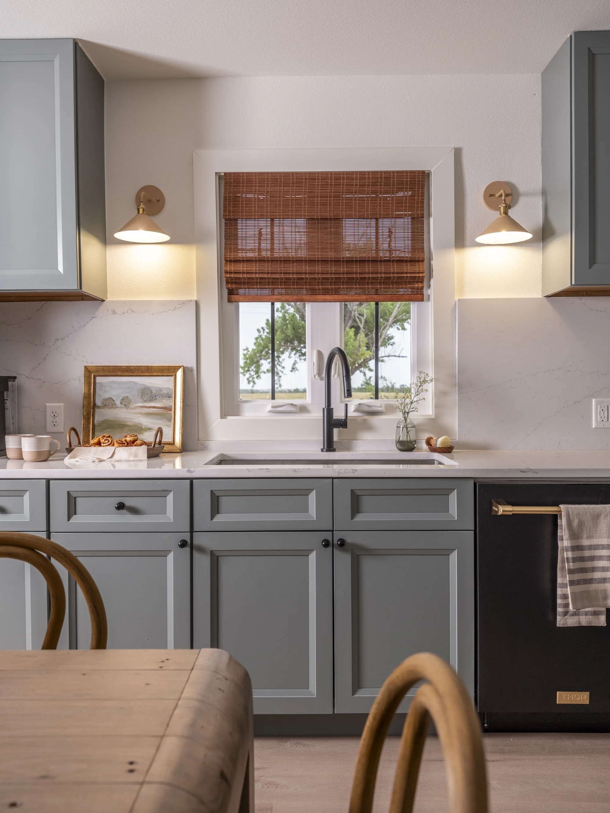 A modern kitchen with light gray cabinets, white marble countertops, a window with bamboo blinds, and a black Thor refrigerator. There are pendant lights on either side of the window and a wooden dining table and chair in the foreground.