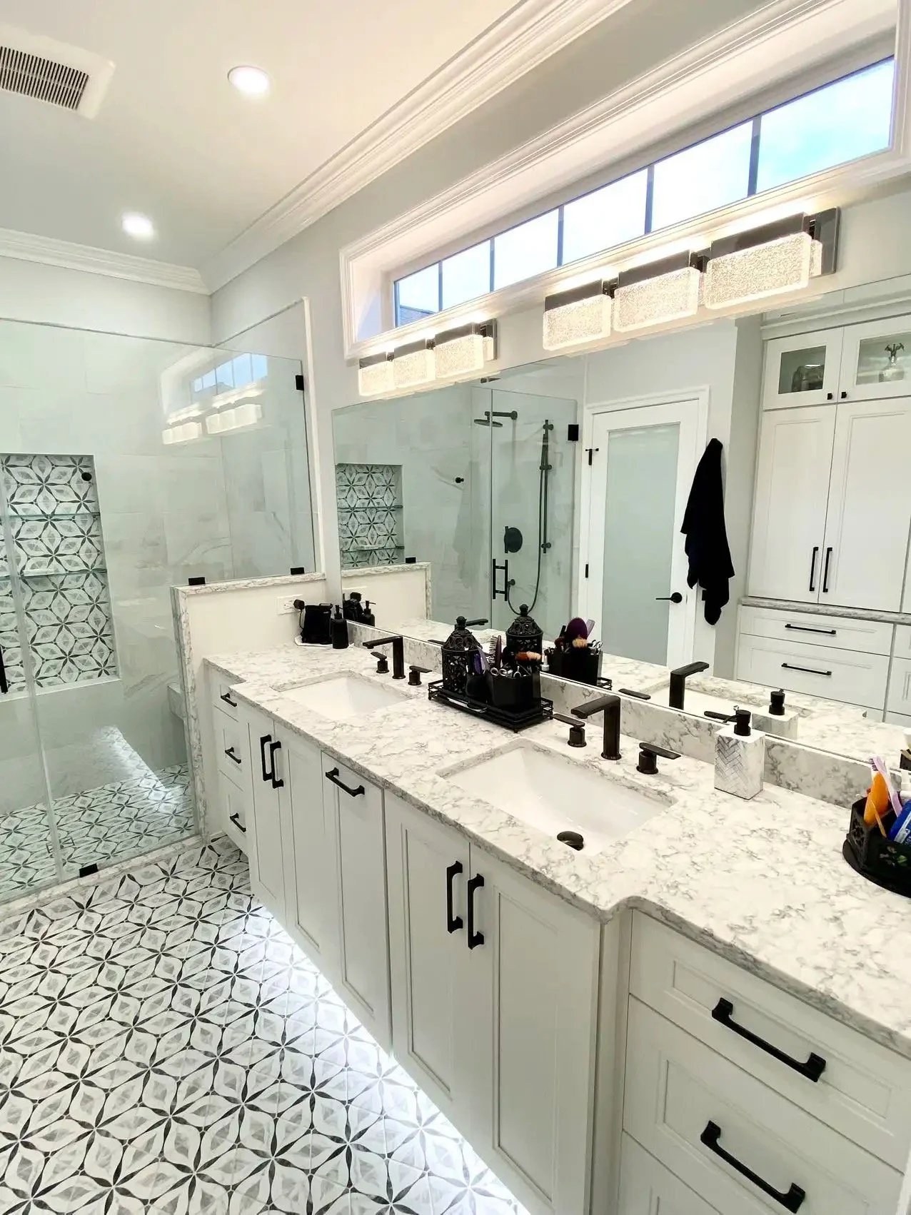 A modern bathroom with a white marble countertop vanity, black hardware, a large mirror, a glass-enclosed walk-in shower with patterned tiles, and a high transom window letting in natural light.