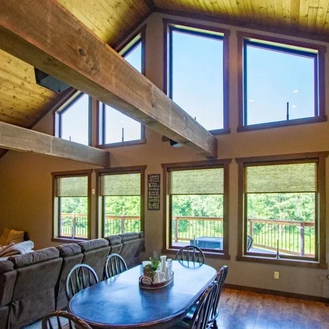 Interior of a house with high vaulted ceiling, large triangular windows, and multiple smaller windows, wooden beams, a dining table with chairs, and a sectional sofa in a bright room with a view of green trees outside.