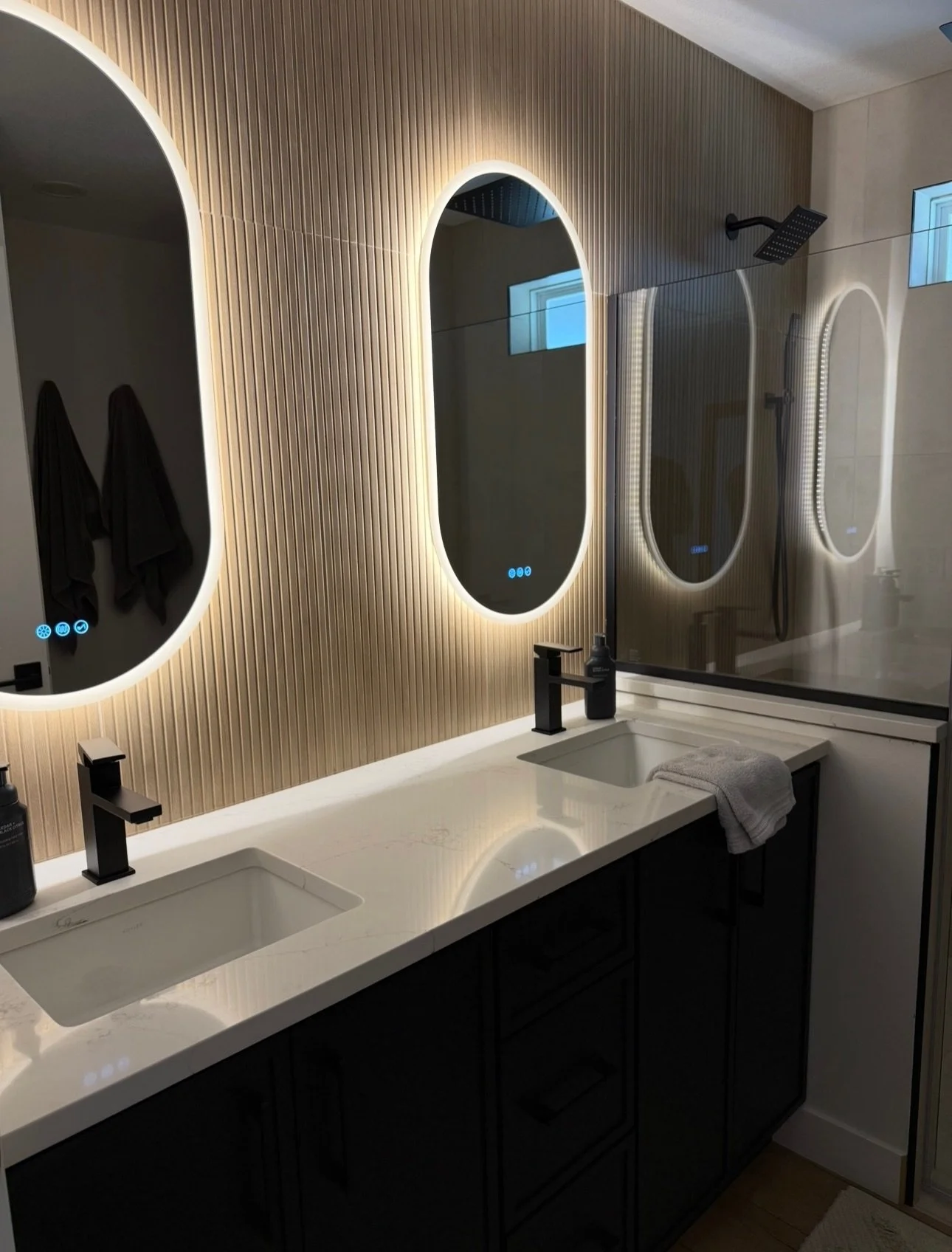 Modern bathroom with dual sinks, black faucets, and backlit mirrors mounted on a textured beige wall. A glass shower enclosure with a rainfall showerhead is visible, along with a window providing natural light. Towel and soap on the countertop.
