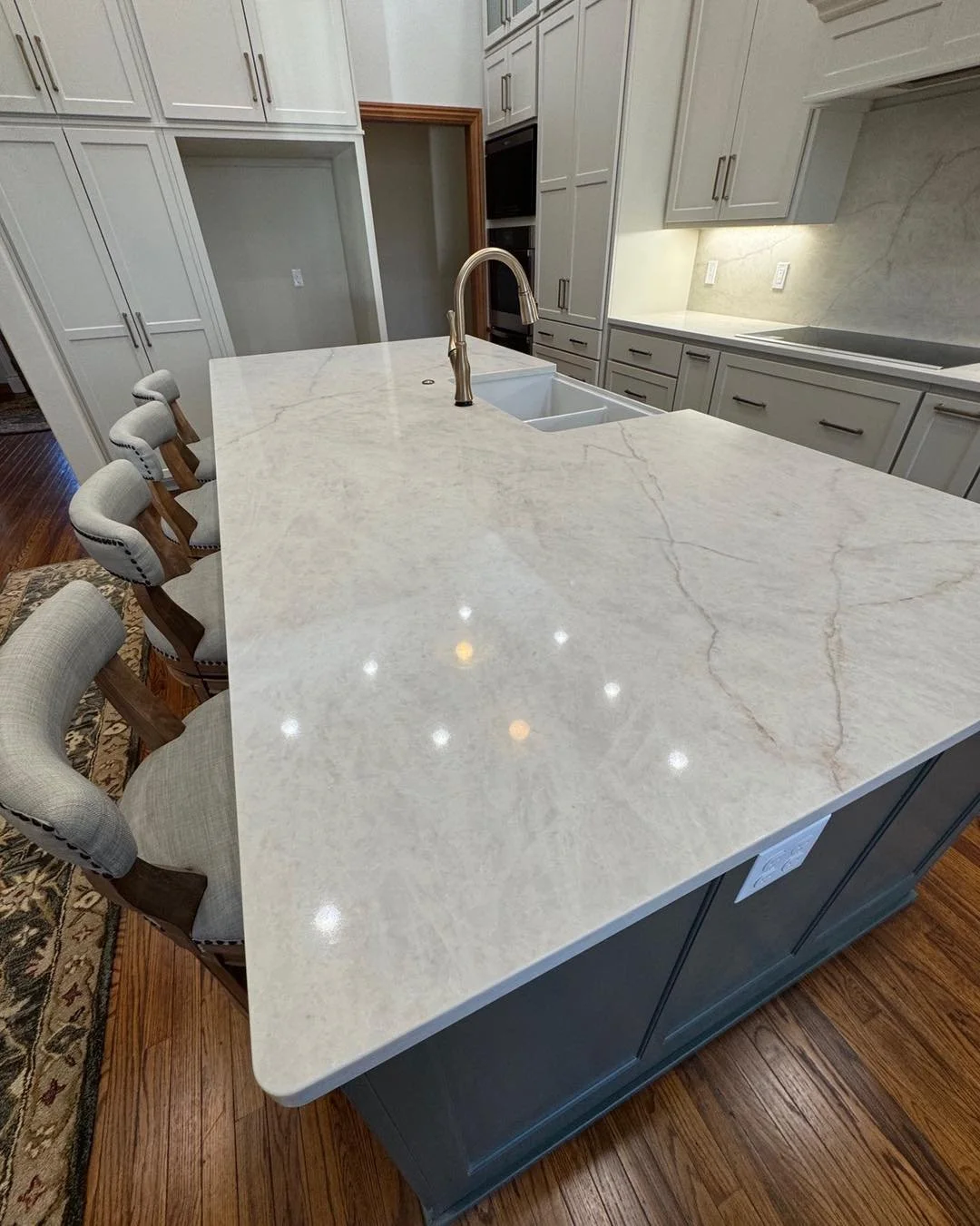 A modern kitchen with a large white marble island, a built-in sink with a modern faucet, and gray cabinets. There are four upholstered chairs with wooden frames along one side of the island. The kitchen has a hardwood floor and under-cabinet lighting.