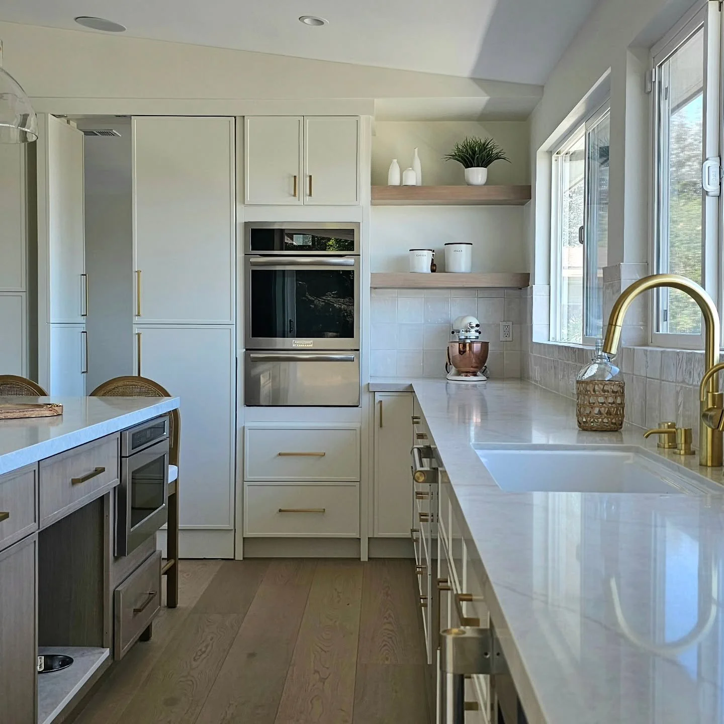Modern kitchen with white cabinets, gold handles, a white marble countertop, a gold faucet, and a window with sunlight. Decorative vases and plants on open shelves, stainless steel oven, and wooden chairs.