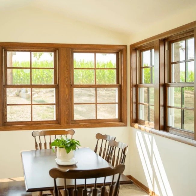 A cozy dining nook with a wooden table, four wooden chairs, a potted plant, and large corner windows showing greenery outside.