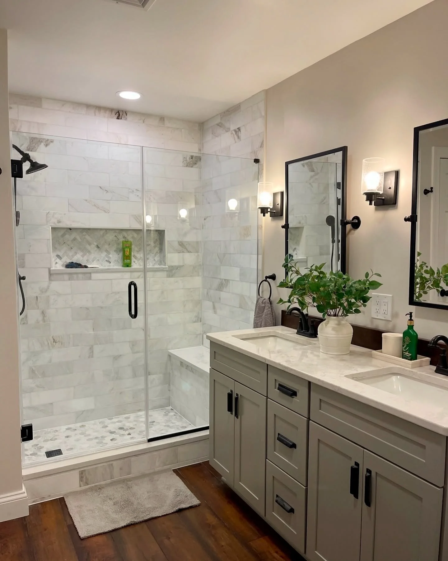 Modern bathroom with a glass shower enclosure, white marble tiles, a beige vanity with black handles, two mirrors, wall-mounted lights, a white vase with green plants, and wood flooring.