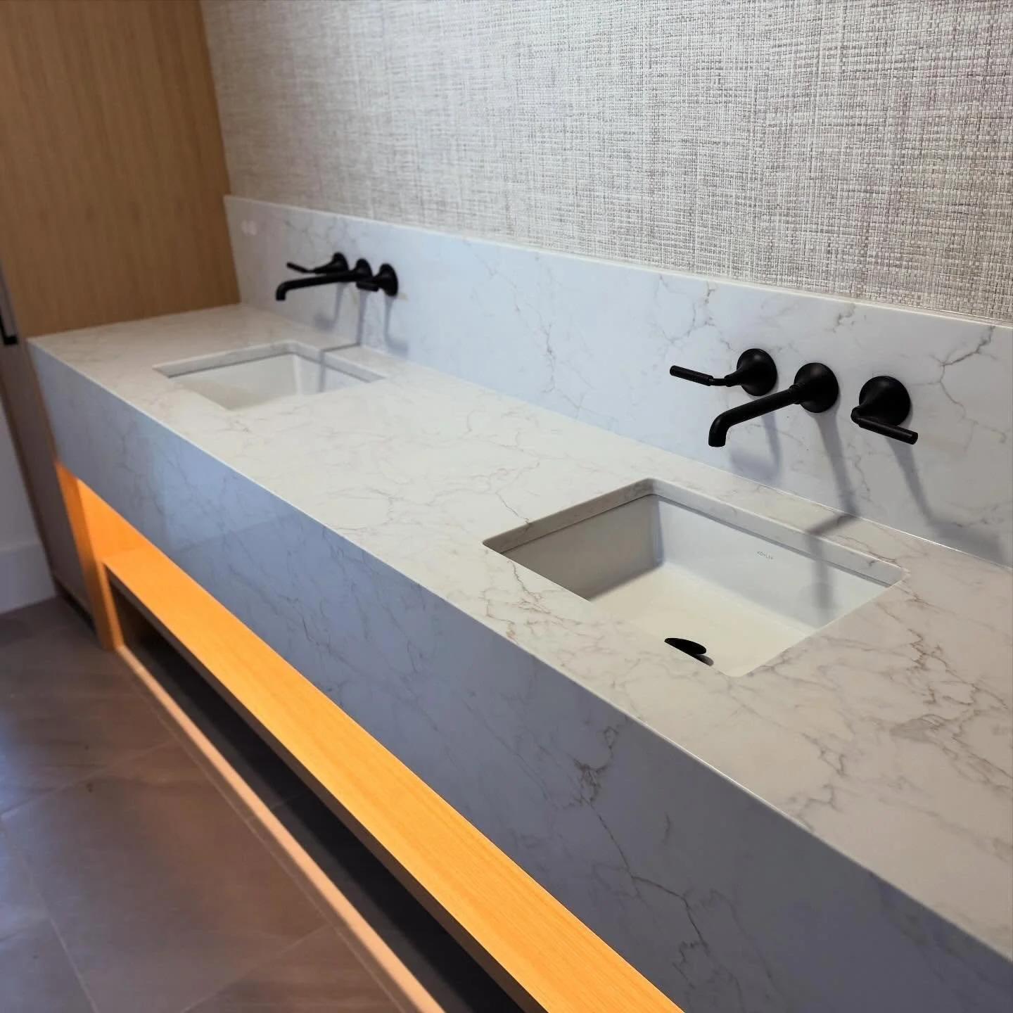 Modern bathroom vanity with a white marble countertop, two square sinks, black wall-mounted faucets, and a wooden illuminated display at the bottom.