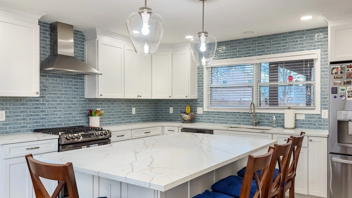 Modern kitchen with white cabinets, a large white marble island, stainless steel appliances, blue tile backsplash, and wooden bar stools.