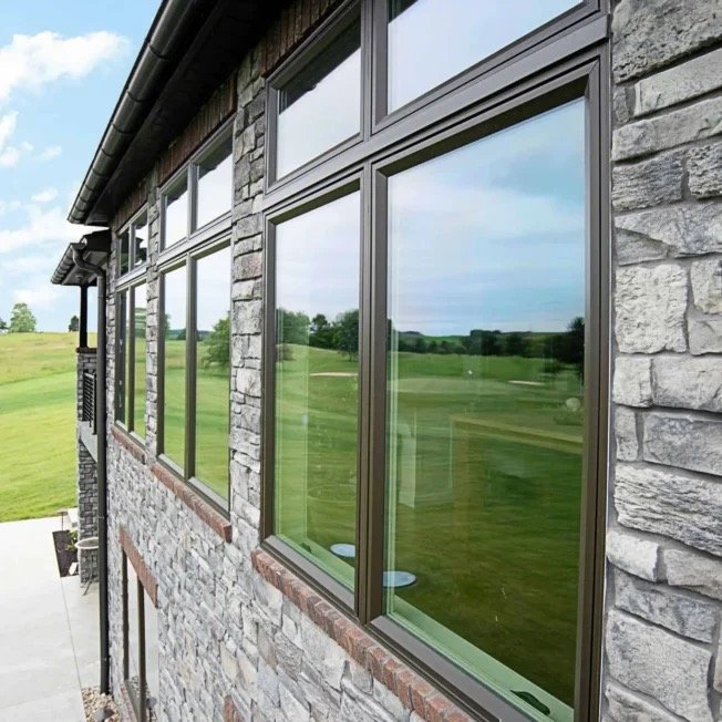 Close-up of a stone house exterior with multiple large windows reflecting a green landscape and blue sky.