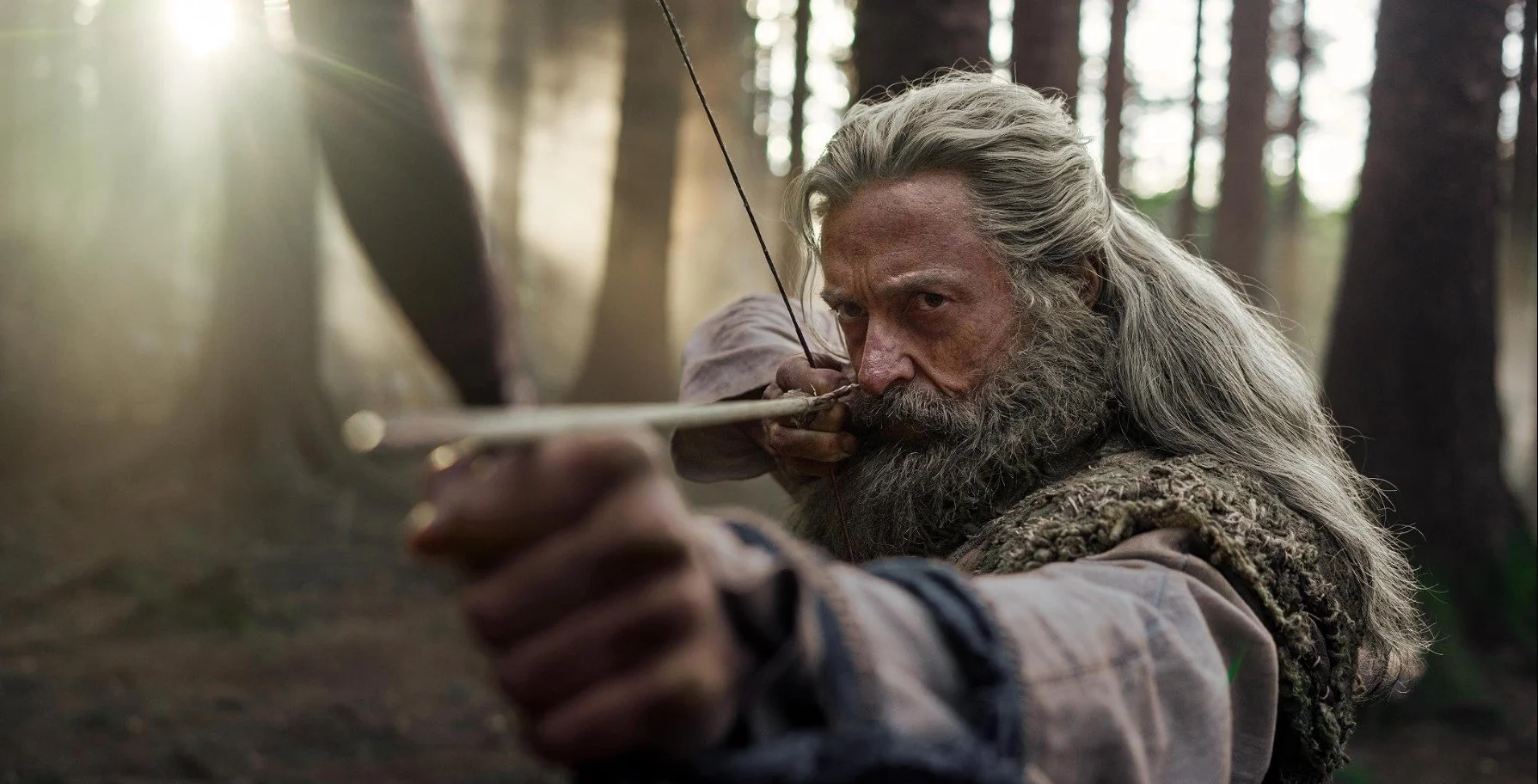 A man with long gray hair and beard, dressed in medieval-style clothing, aiming a bow and arrow in a forest.