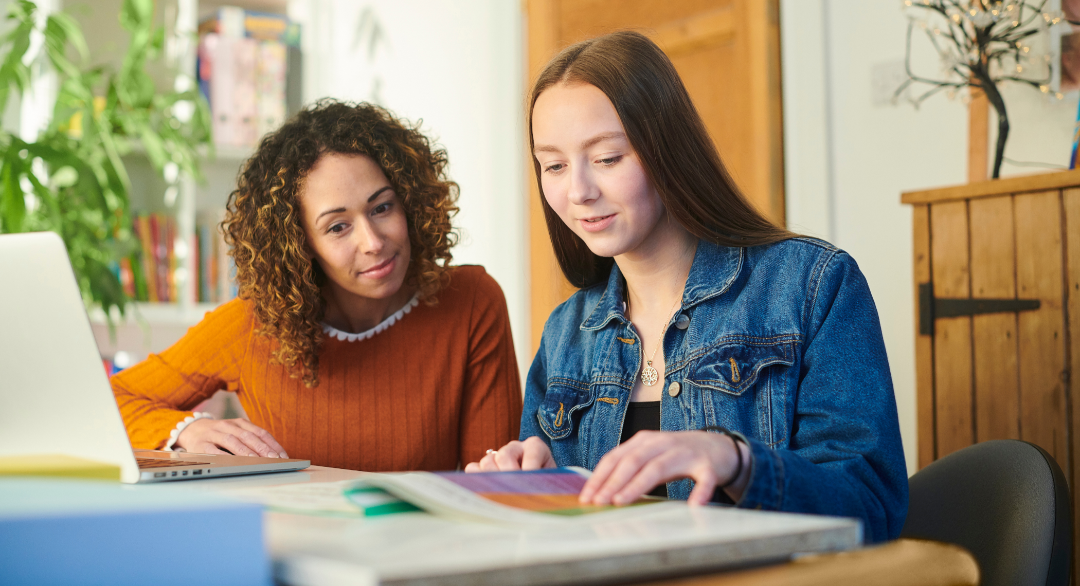 Two women sit at a desk, looking at an open book or magazine. One woman with long brown hair wearing a denim jacket is showing something to the other woman with curly hair in an orange sweater. There are bookshelves, a potted plant, and a wooden cabinet in the background.