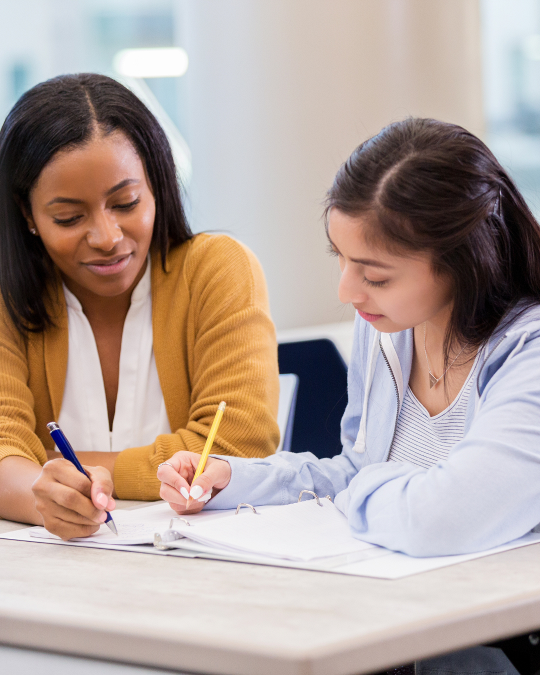 Two women sitting at a table, looking at papers and writing with pens in an indoor setting.