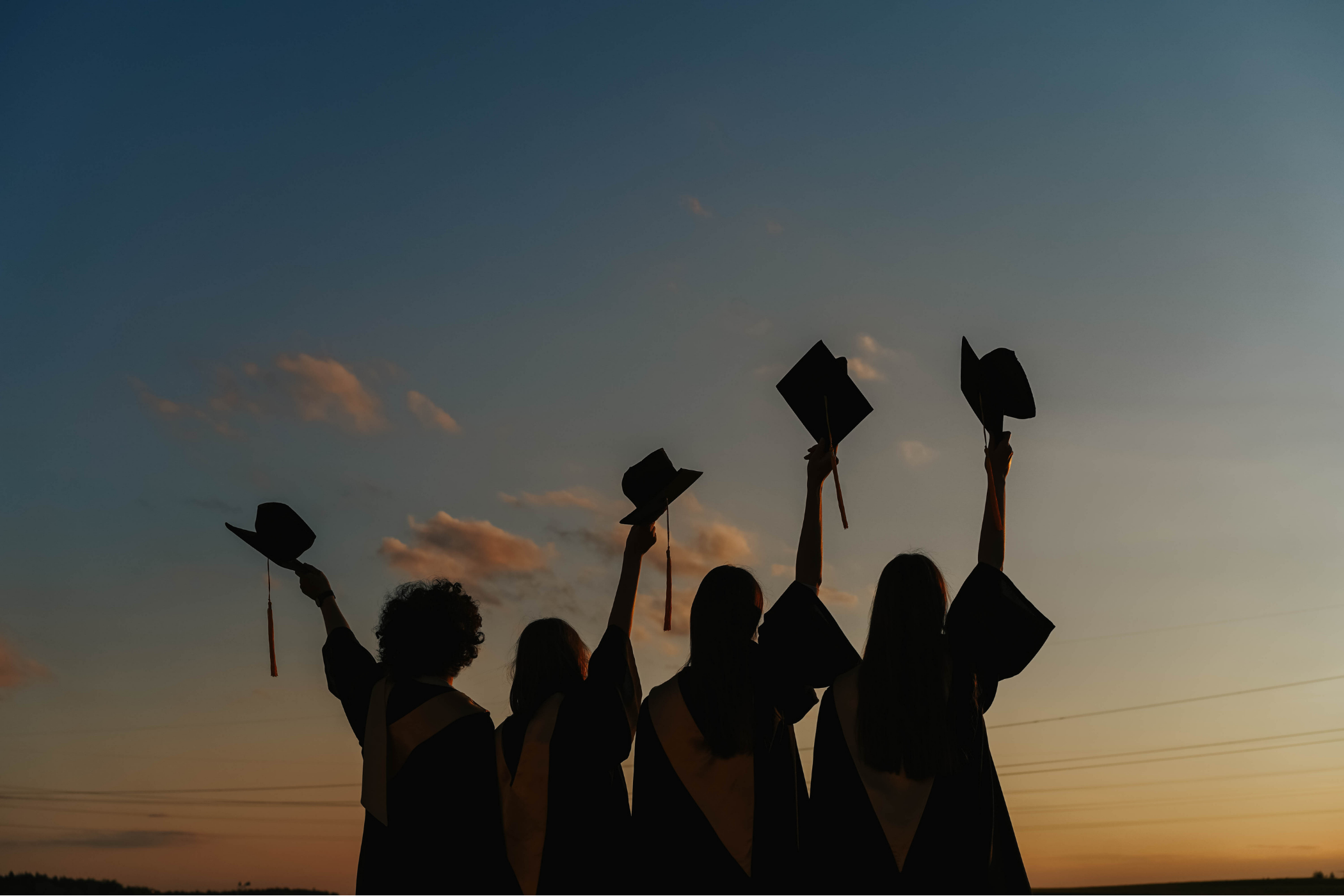Silhouettes of graduates holding graduation caps in the air during sunset.