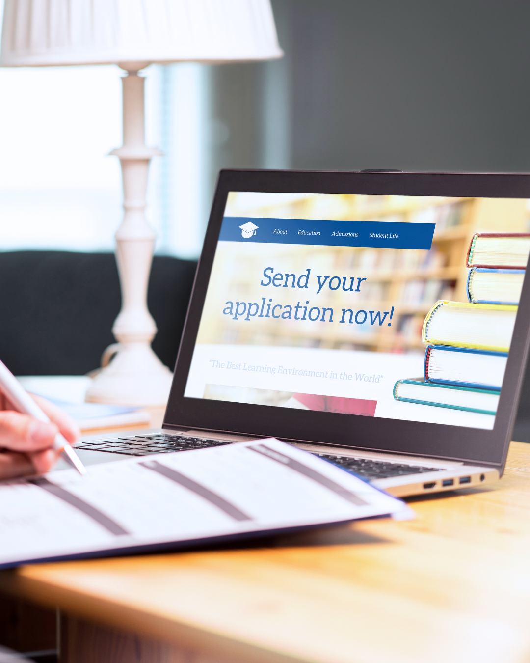 Laptop on a wooden desk displaying an online application form with the headline 'Send your application now!' and a stack of books next to it.