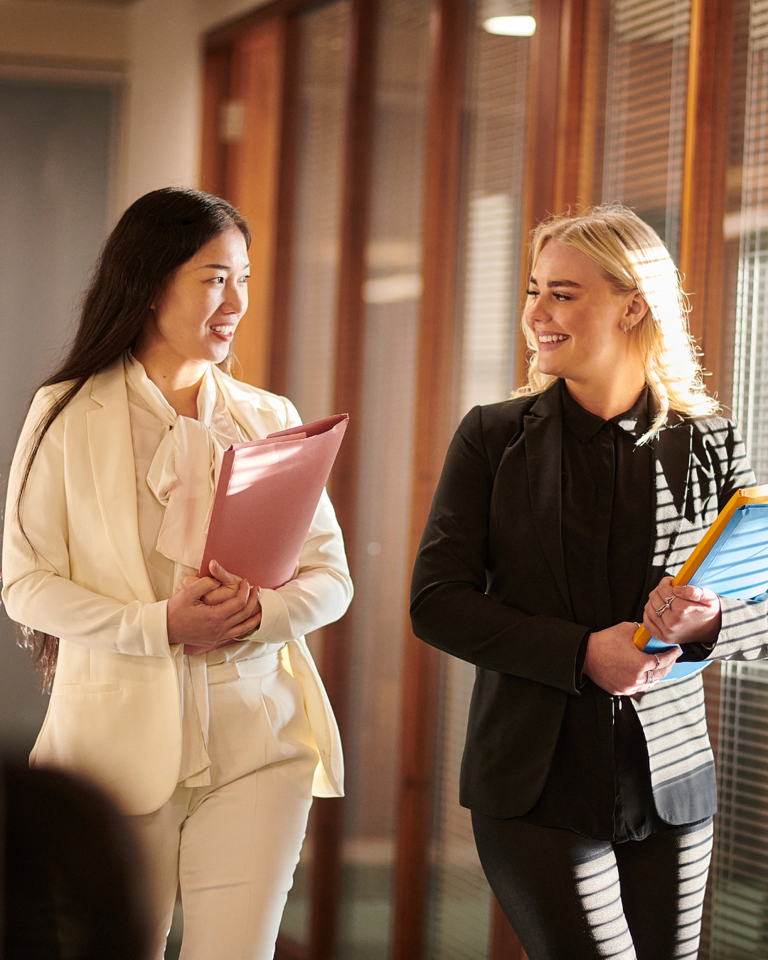 Two women smiling and walking together, carrying folders in a well-lit office corridor with wooden paneling.