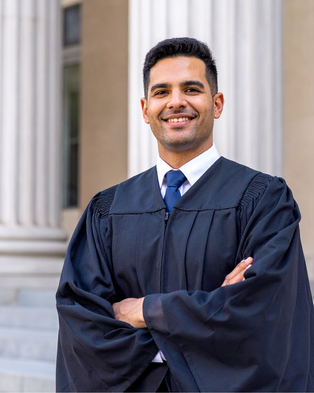 A man in a graduation cap and gown smiling in front of a building with columns.