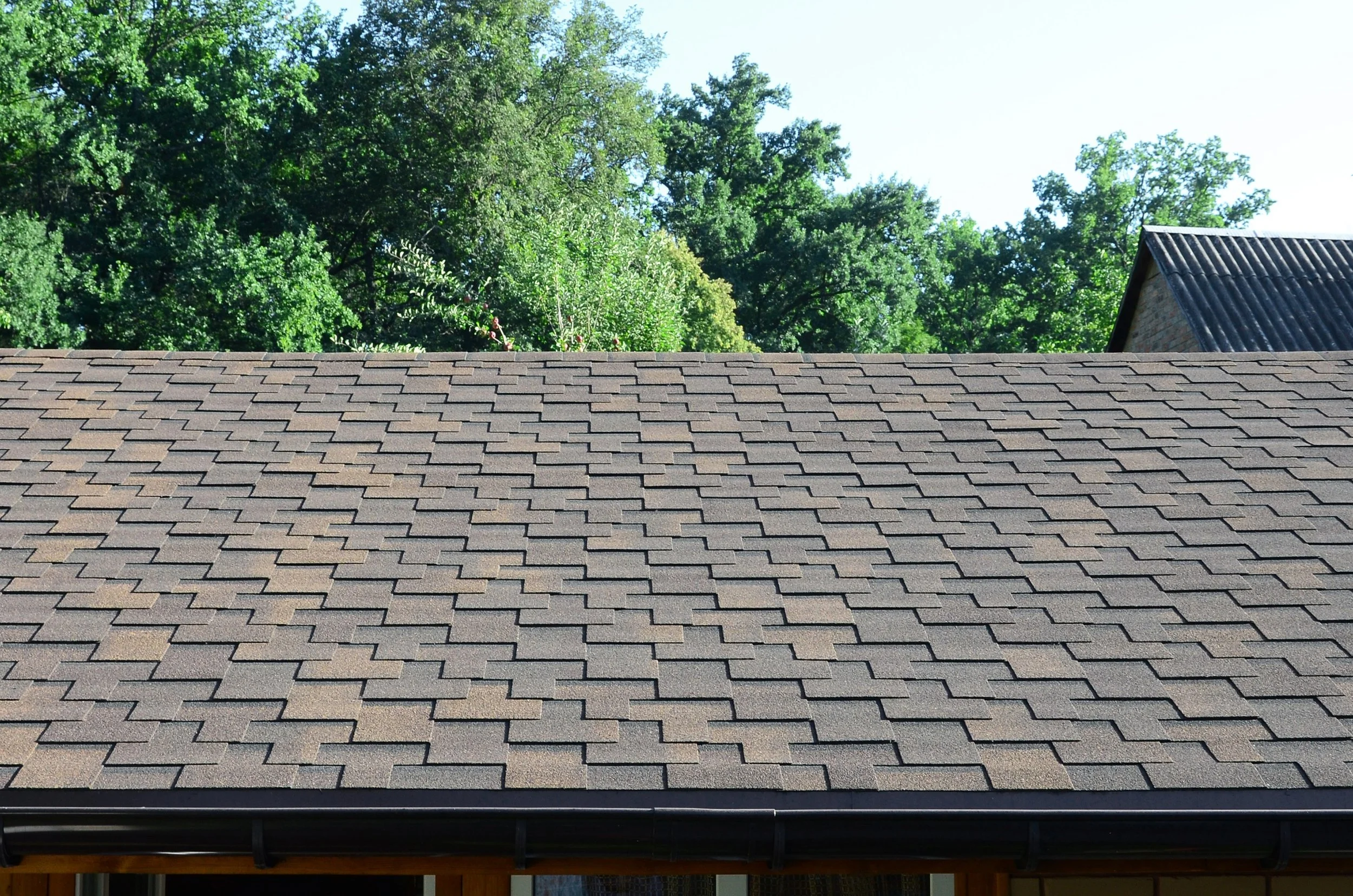 Roof with gray shingles and a rain gutter, with green leafy trees and a partially cloudy sky in the background.