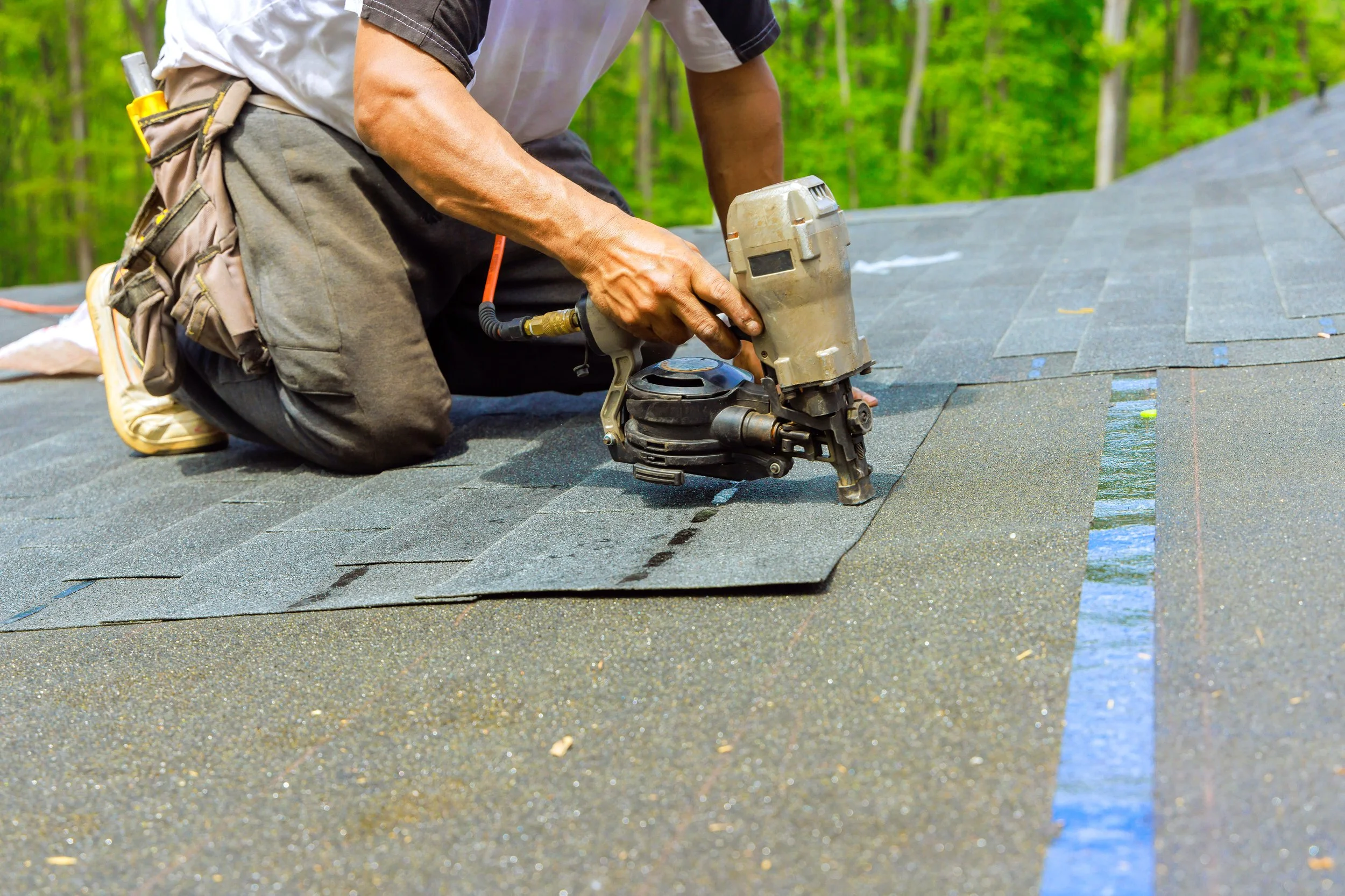 A person installing asphalt shingles on a roof with a nail gun, wearing a tool belt, in a wooded area.