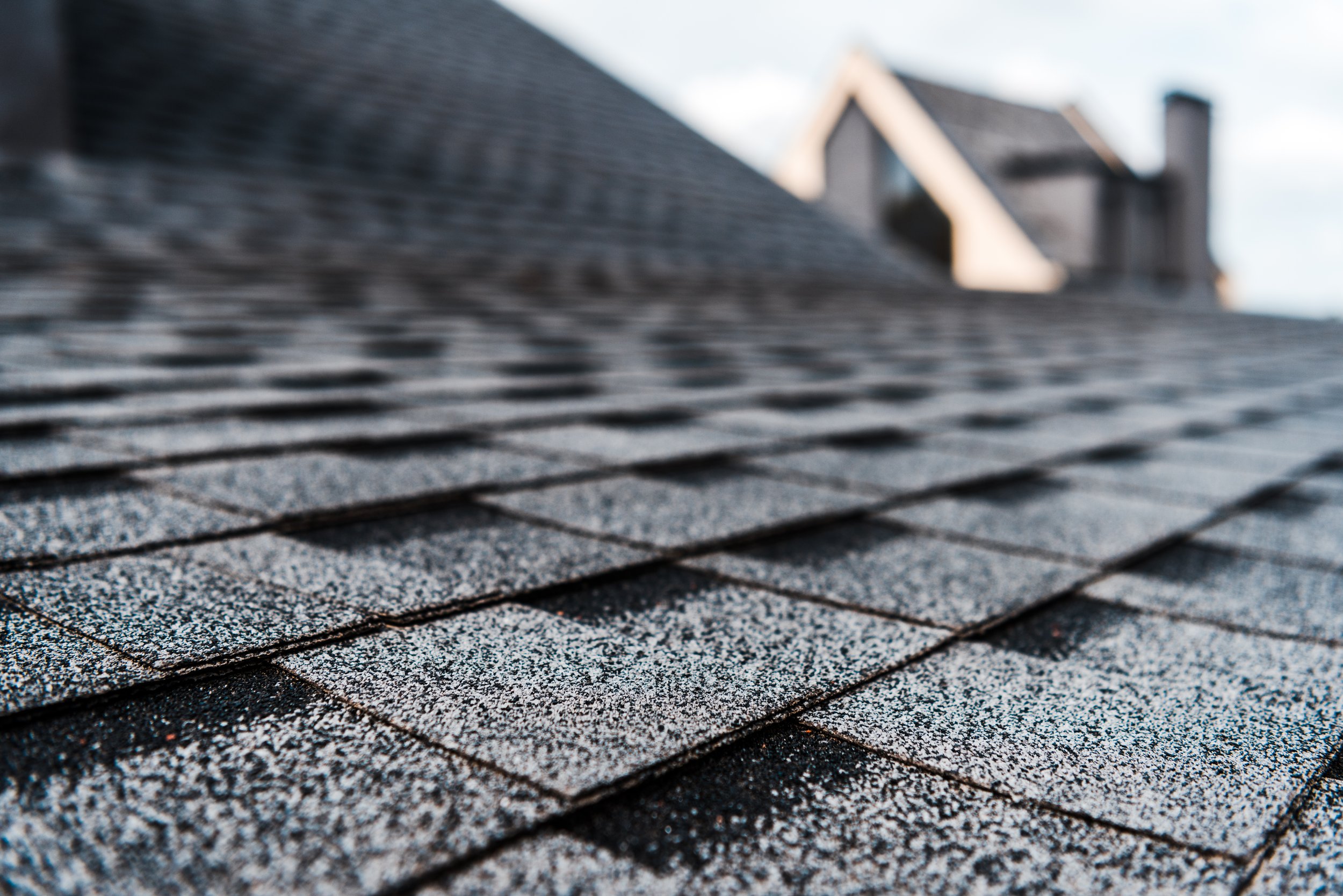 Close-up of asphalt shingles on a roof, with a house and chimney in the background, under a cloudy sky.