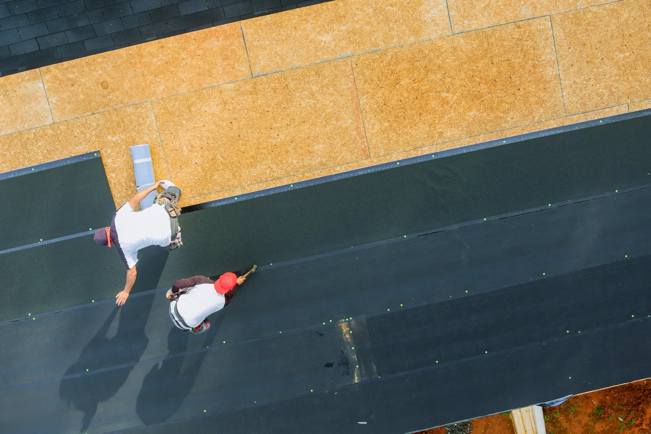 Two construction workers installing black roofing material on a sloped roof, with a section of the roof covered in orange underlayment.