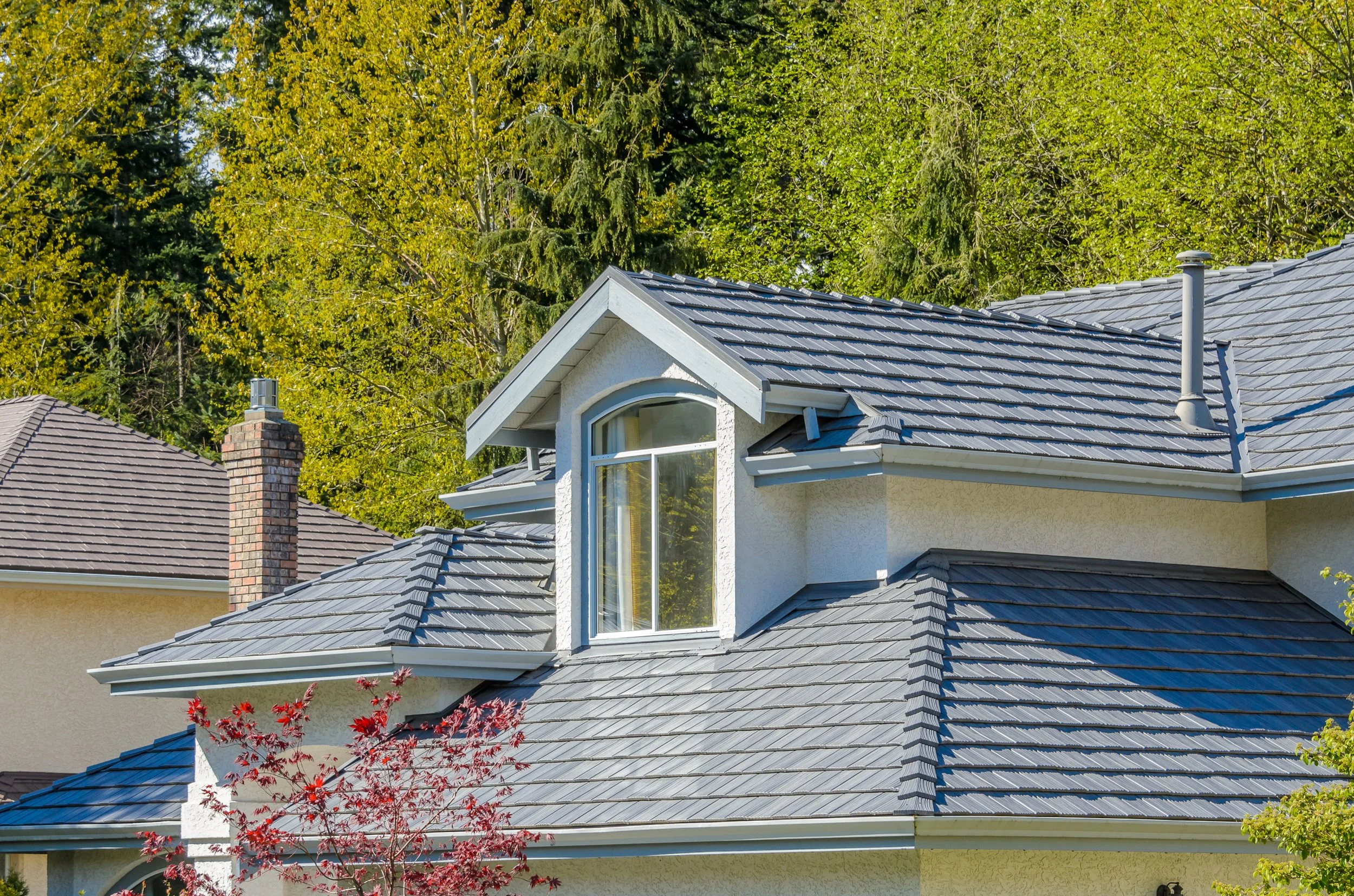 Closer view of house rooftops with gray shingles, featuring a prominent gable window and chimney, surrounded by green trees.