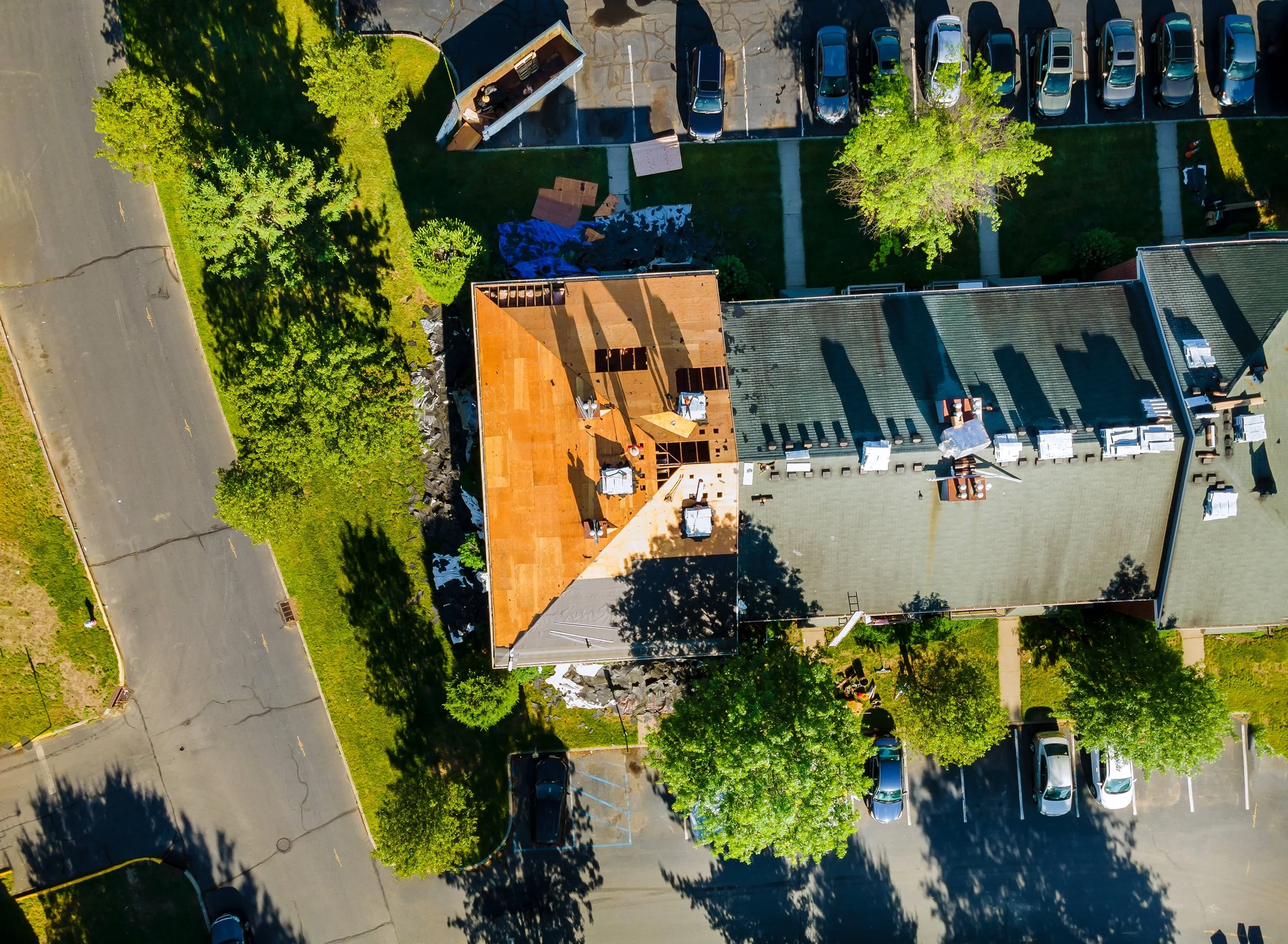 An aerial view of a suburban neighborhood showing a house under construction, a parking lot with several cars, trees, and a paved street.