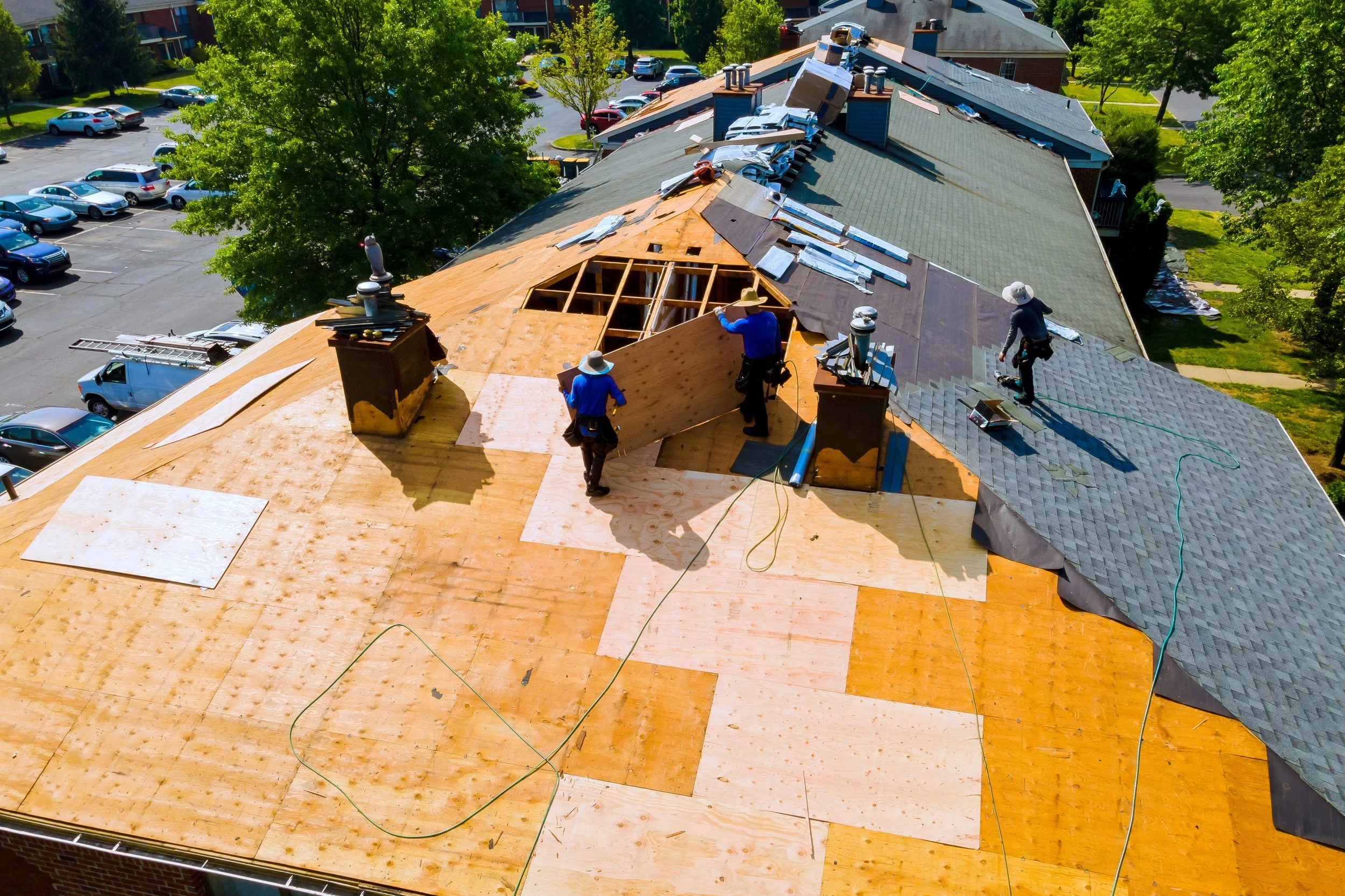 Construction workers are working on the roof of a building, installing or repairing shingles. The roof is partly covered with new shingles, and parts are still under construction with exposed plywood. There are multiple workers wearing hats and safety gear, and various tools and materials scattered across the roof. The building is surrounded by trees, and a parking lot with many cars is nearby.