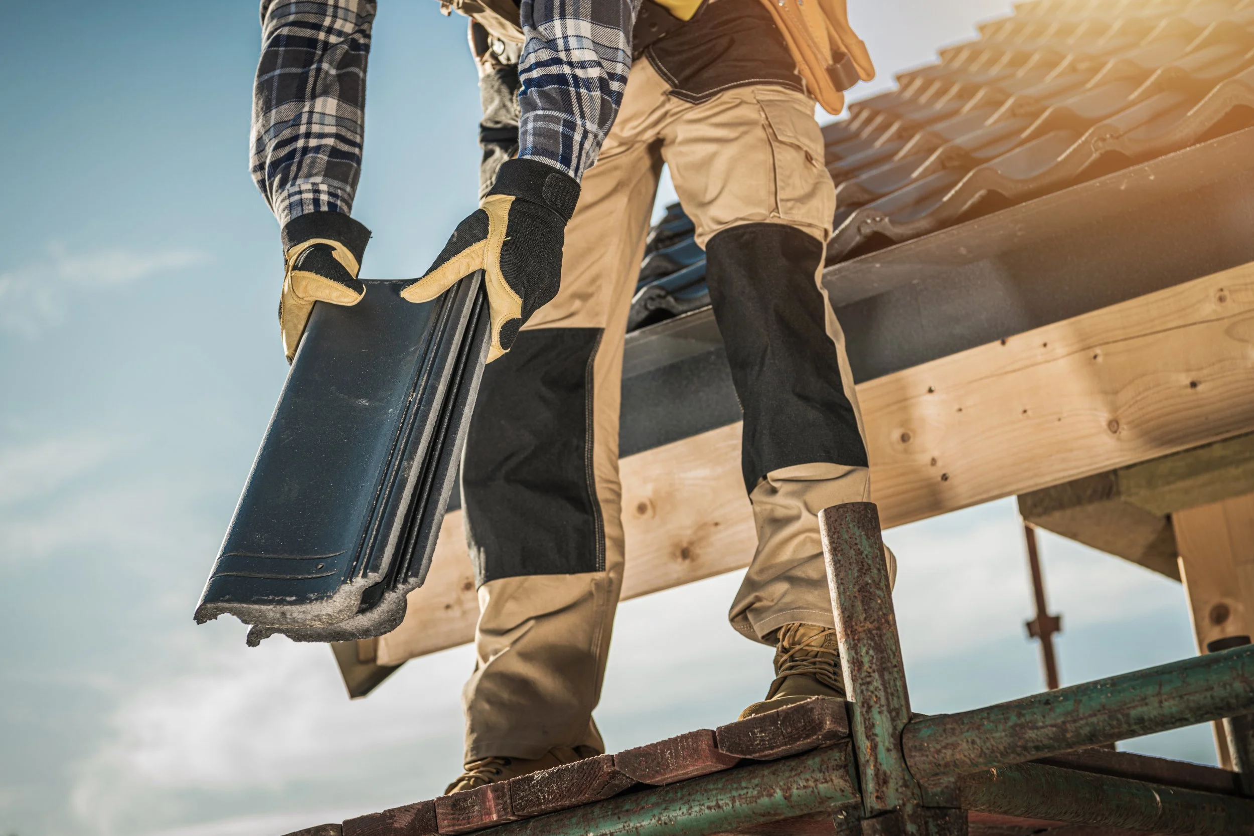 A construction worker standing on scaffolding, wearing gloves and safety gear, working on a building with a blue sky background.