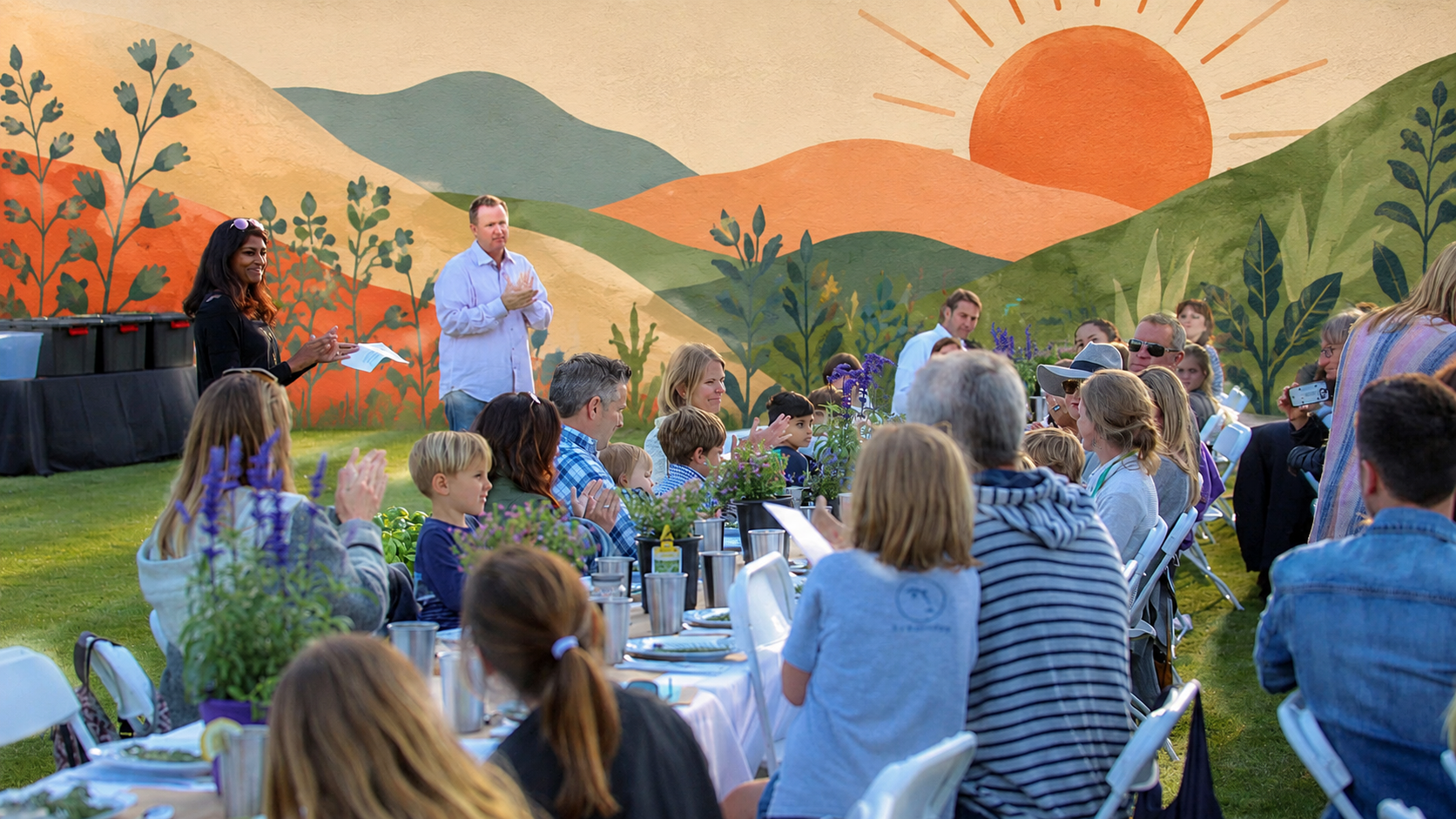 Outdoor gathering with people sitting at long tables, listening to speakers, with a painted mural of mountains and a sun in the background.