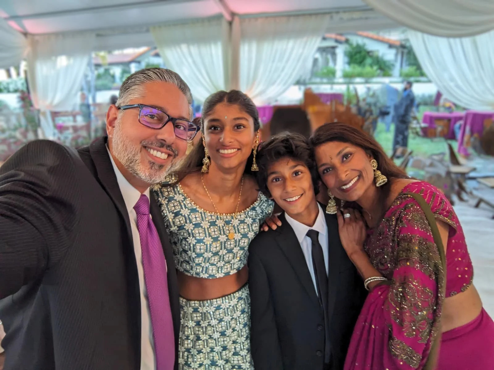 A group of five people smiling and taking a selfie at a celebration or wedding, with a decorated indoor venue in the background.