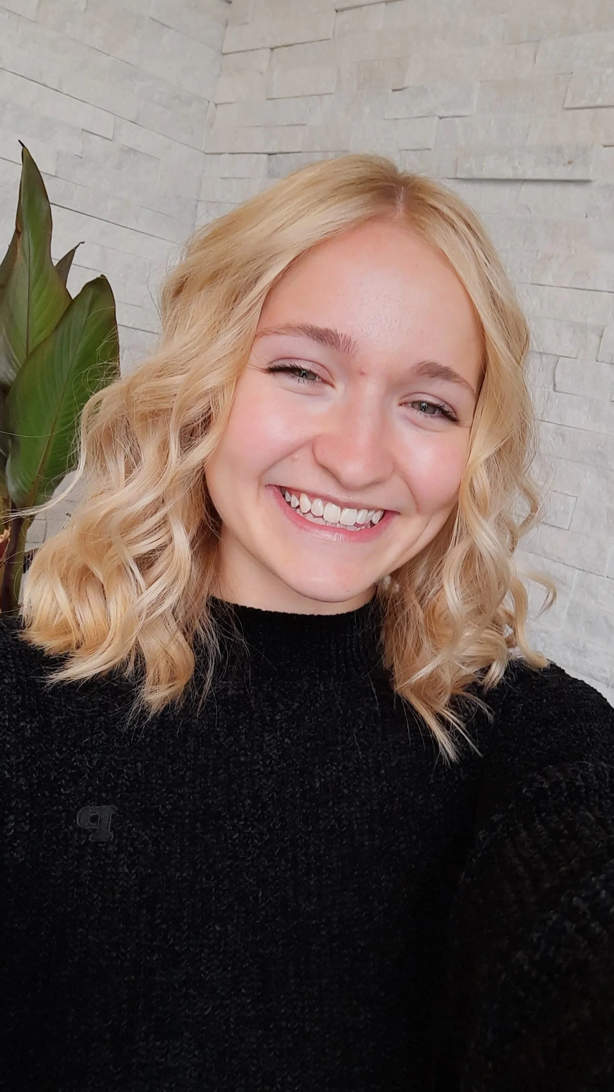 Young woman with wavy blonde hair, smiling, wearing a black sweater, standing against a white brick wall with a large green plant to her left.